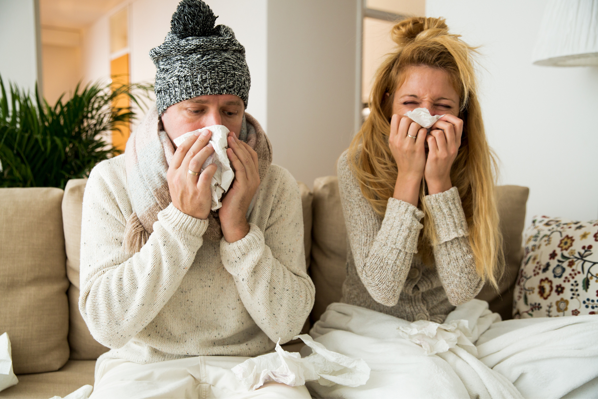 Two people sitting on a couch blowing their noses with tissues, appearing sick or congested