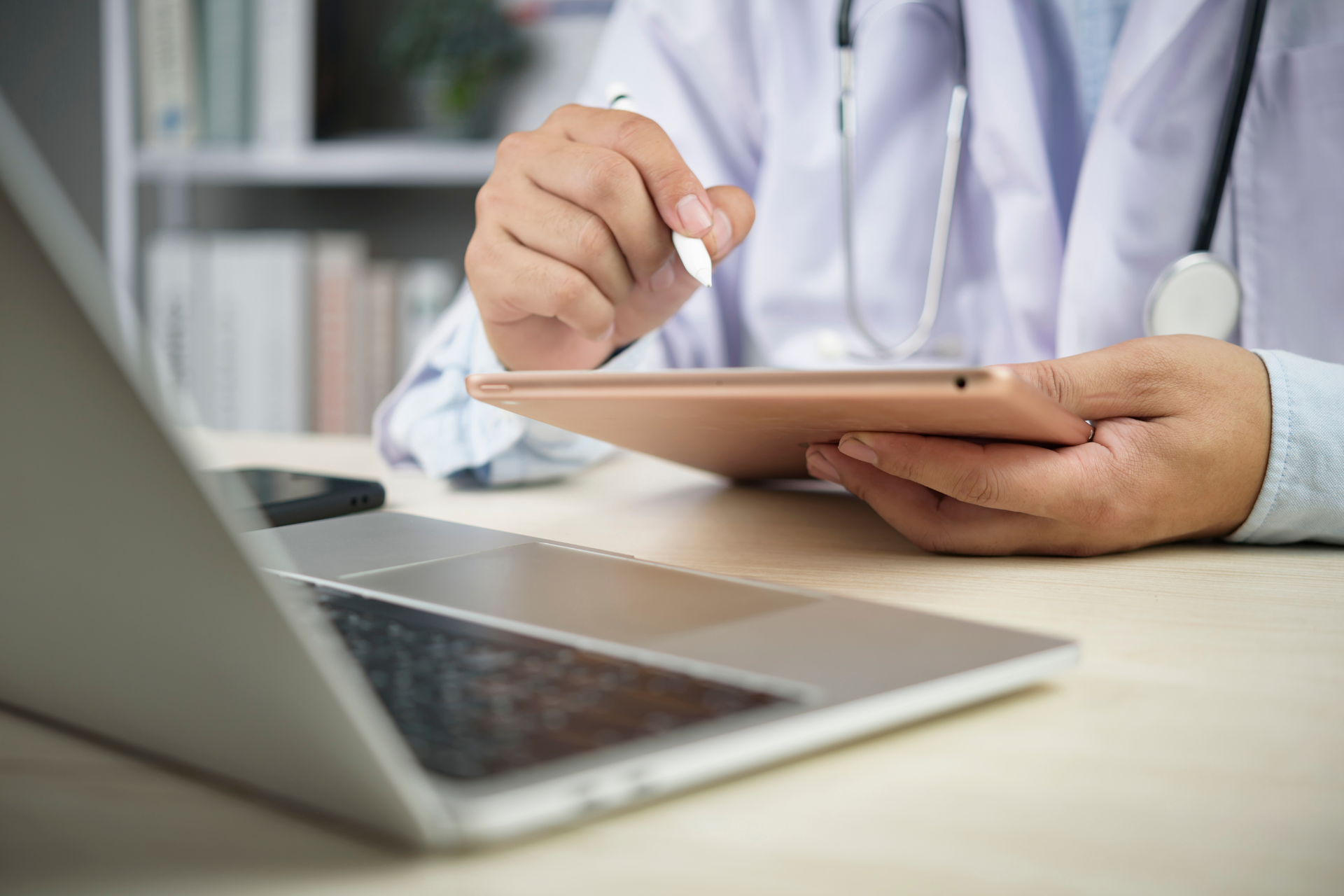 Doctor using a digital tablet with a stylus while sitting at a desk with a laptop, reviewing medical information