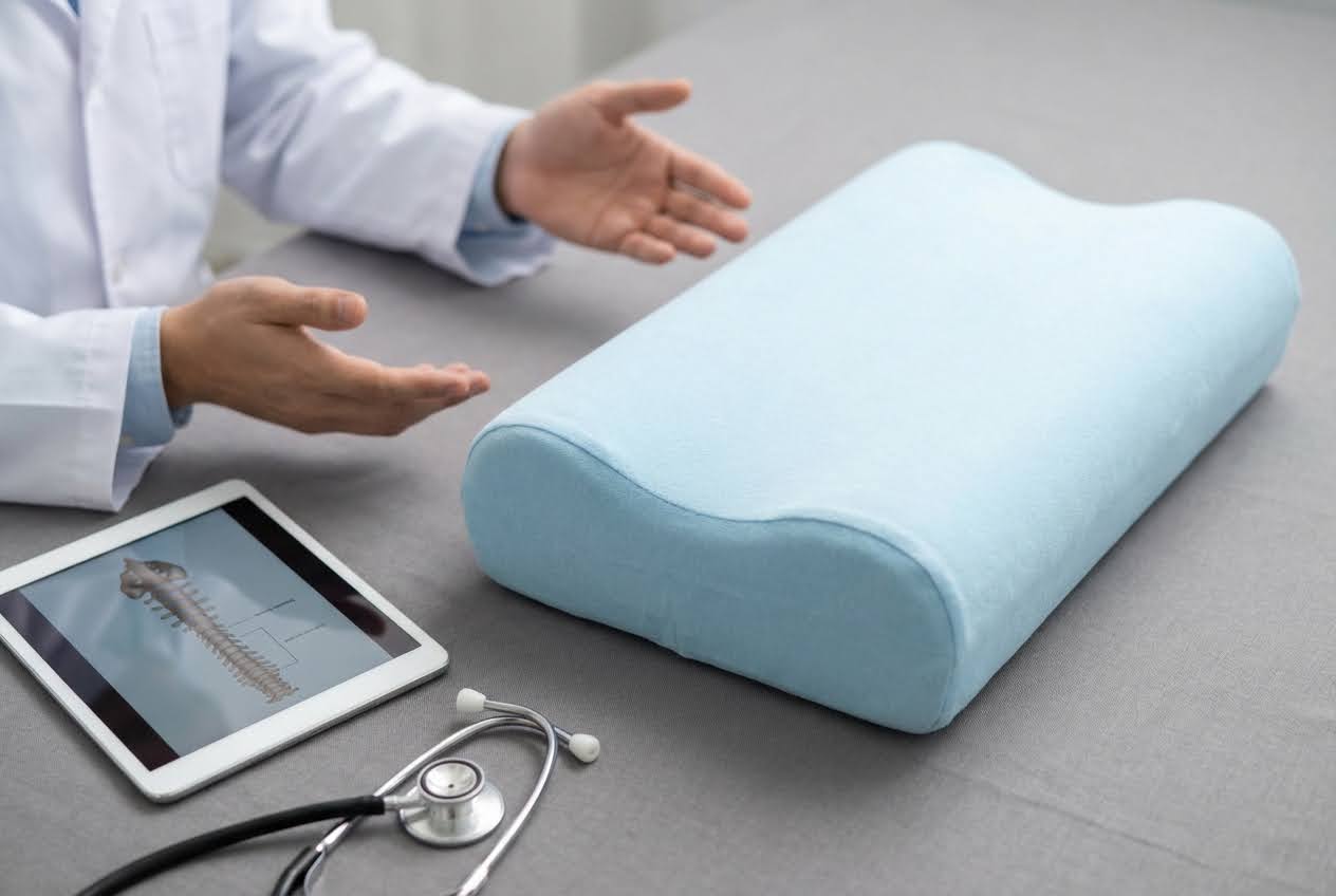 Doctor gesturing towards a light blue cervical pillow, with a tablet showing a spine and a stethoscope on a gray surface.