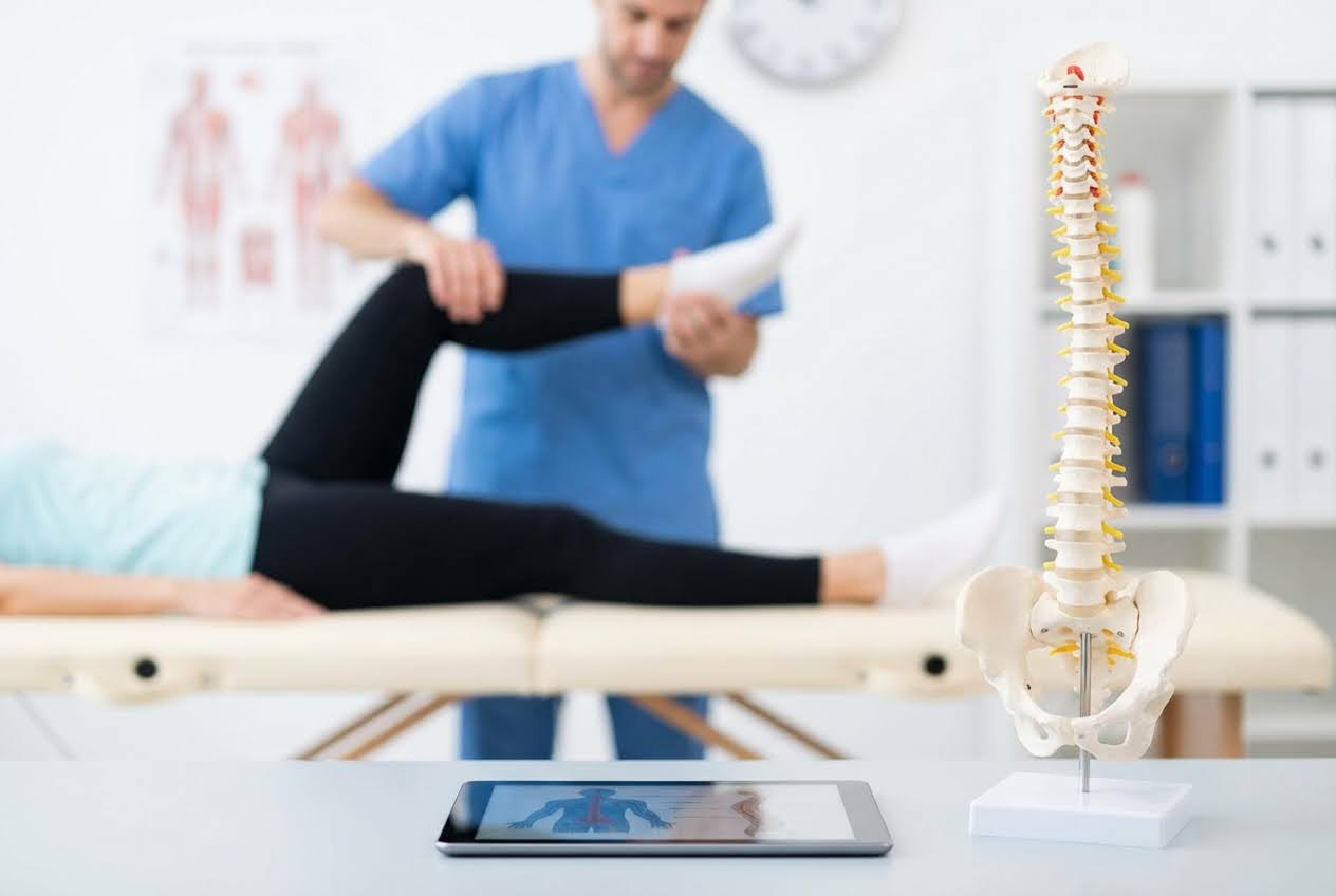 Physiotherapist stretching a patient's leg on an exam table, with a spine model and tablet in the foreground.