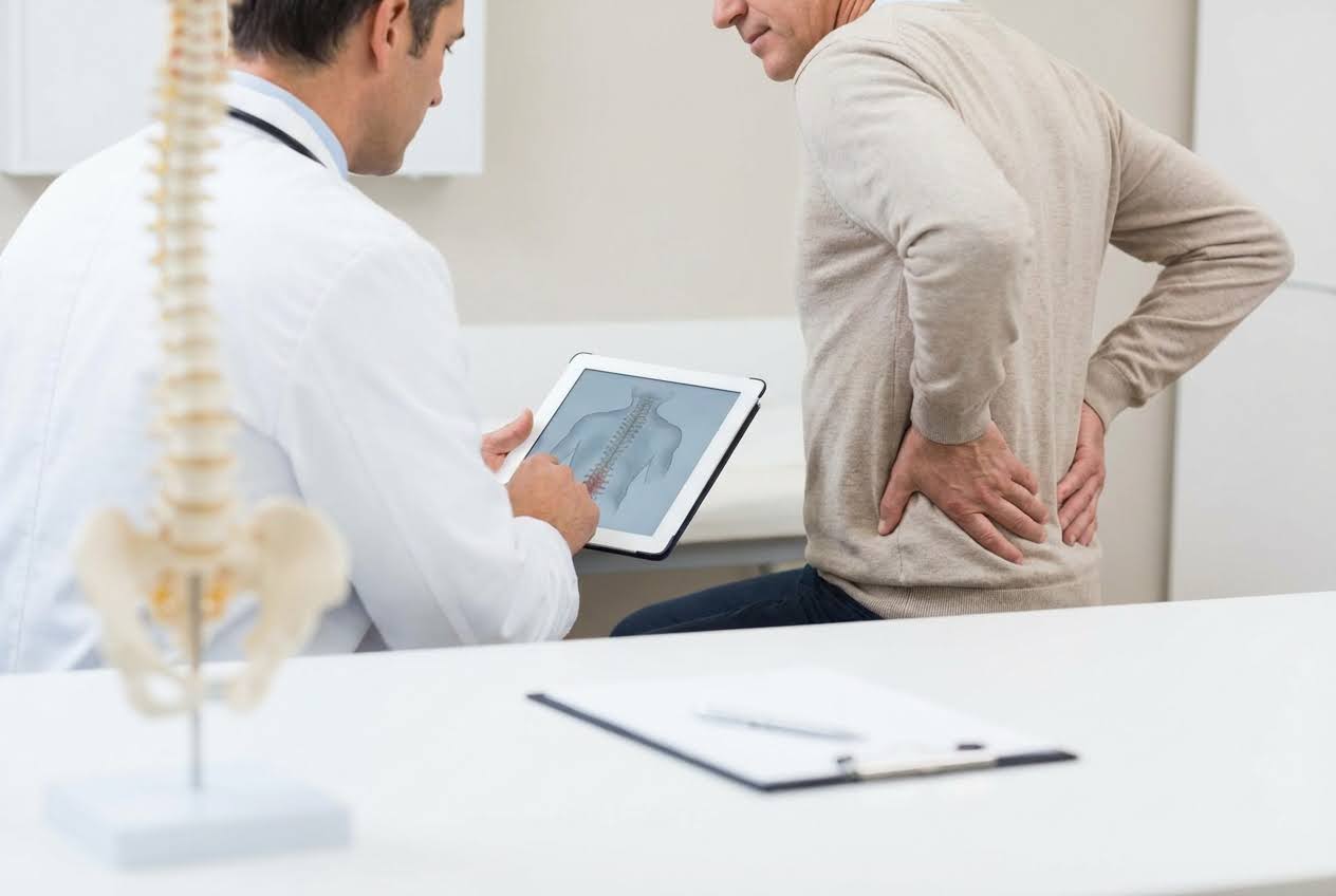 Doctor showing a patient a spine diagram on a tablet, while the patient holds his aching lower back.