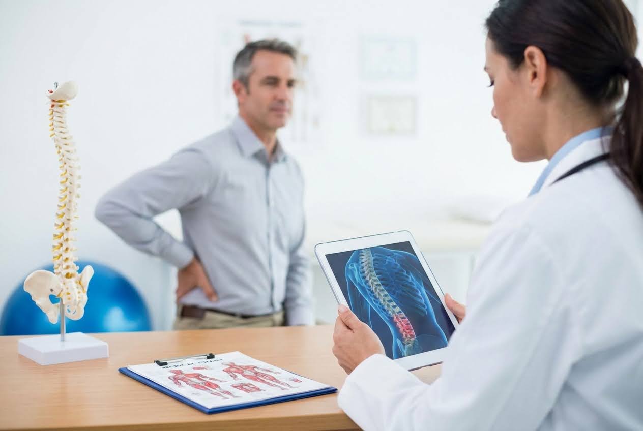 A doctor reviews a spinal scan on a tablet while a man stands nearby holding his lower back, indicating back pain during a medical consultation.