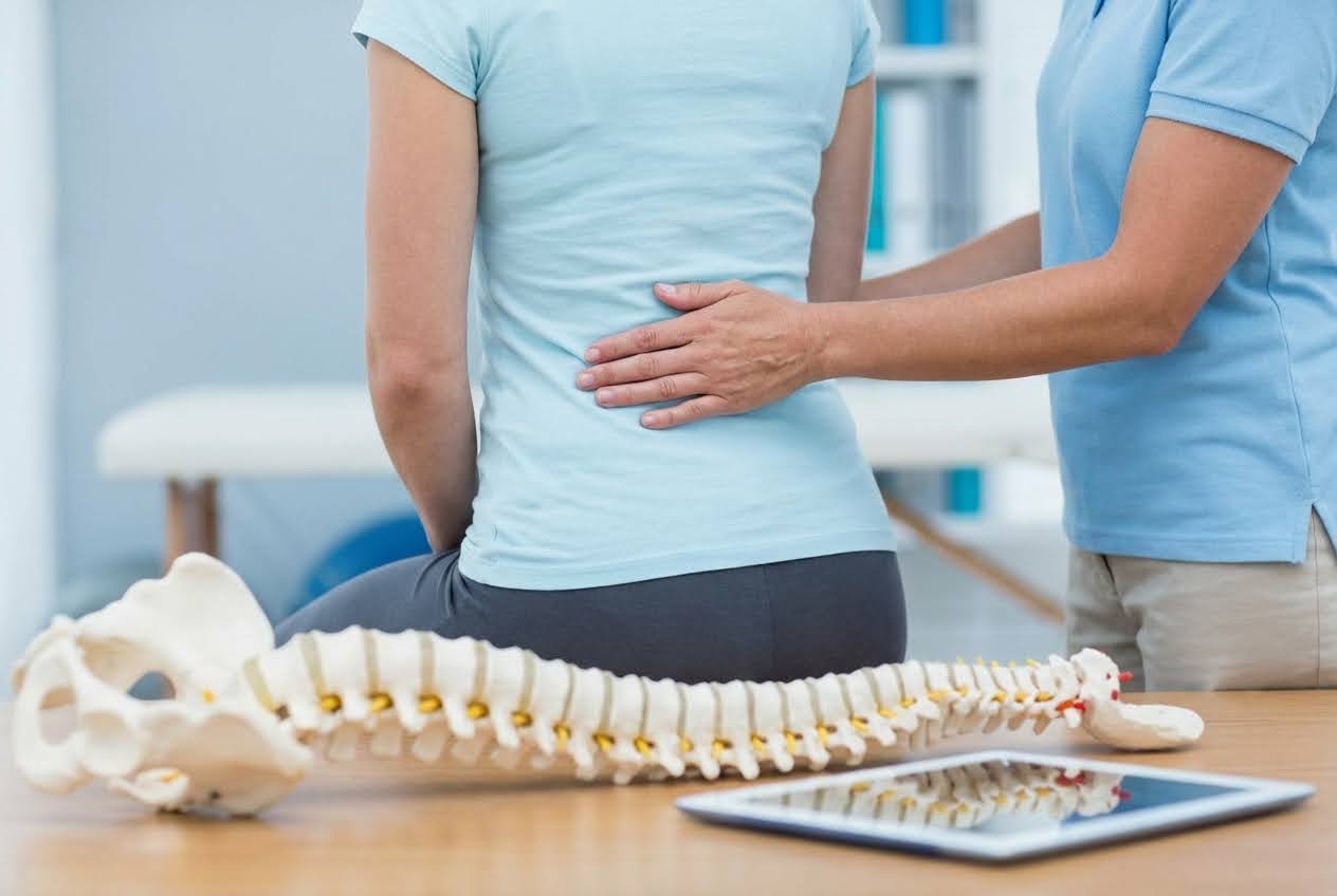 A person in a light blue shirt receiving back pain relief from a physical therapist, with a spine model and tablet on the table.