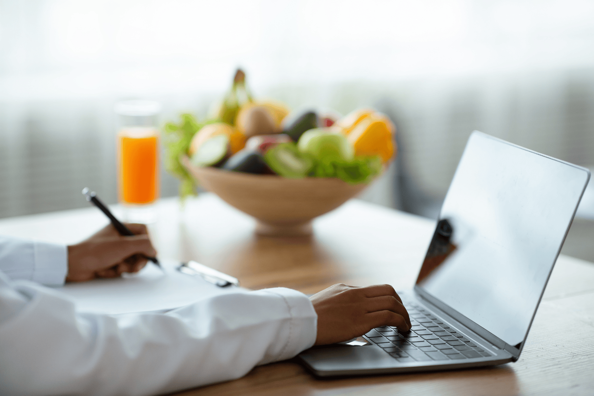 A Nutritionist writing notes while using a laptop with fruits and juice on the table.
