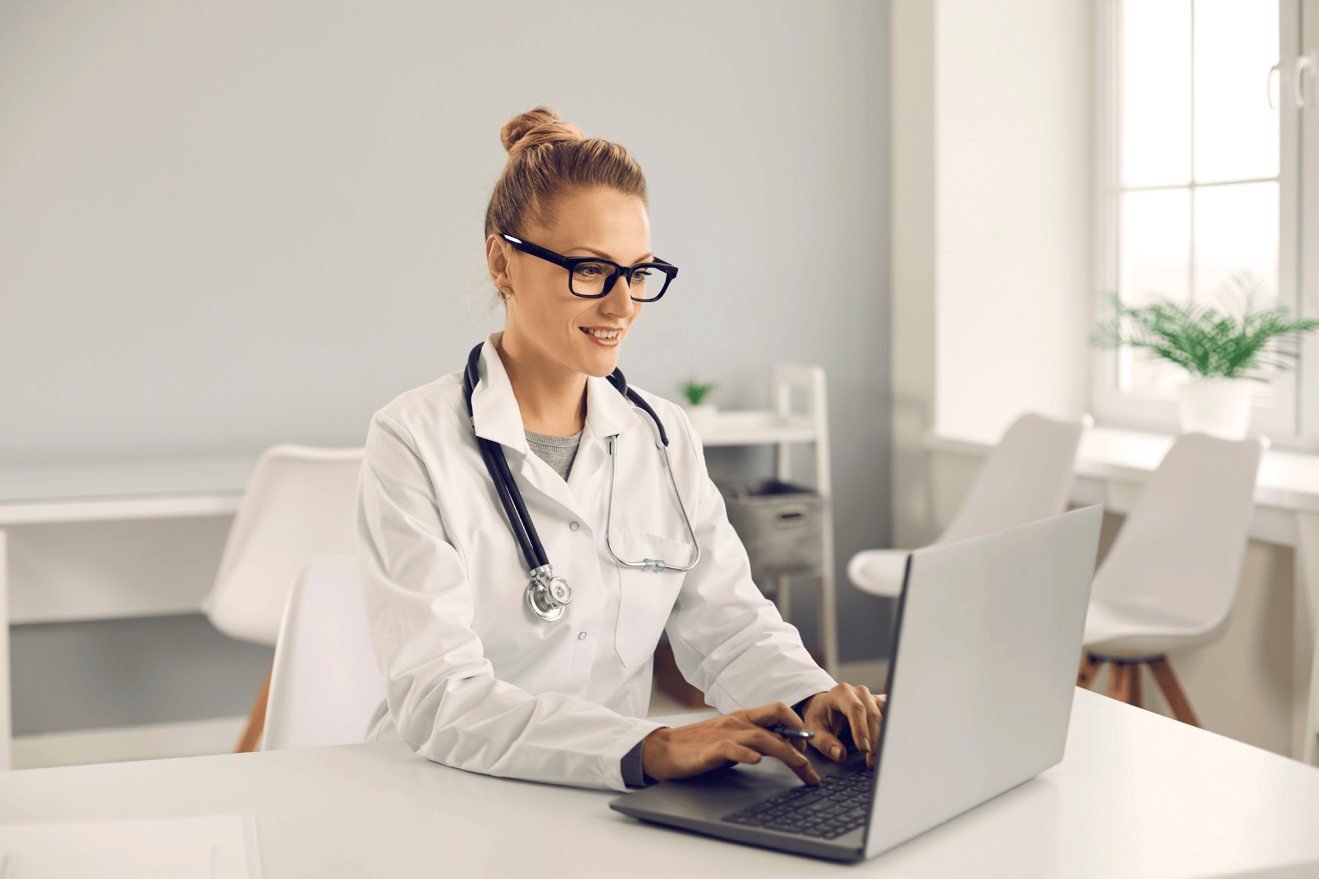 Female doctor with a stethoscope working on a laptop in a bright office.