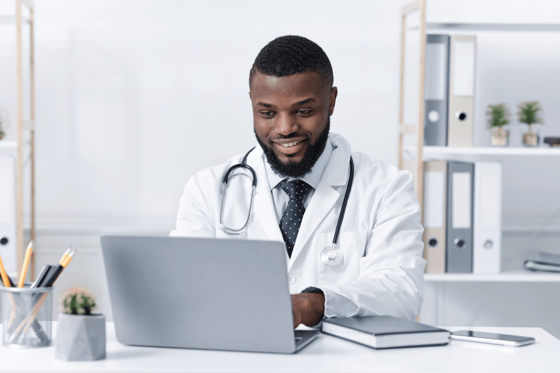 Smiling doctor with a stethoscope working on a laptop in an office.