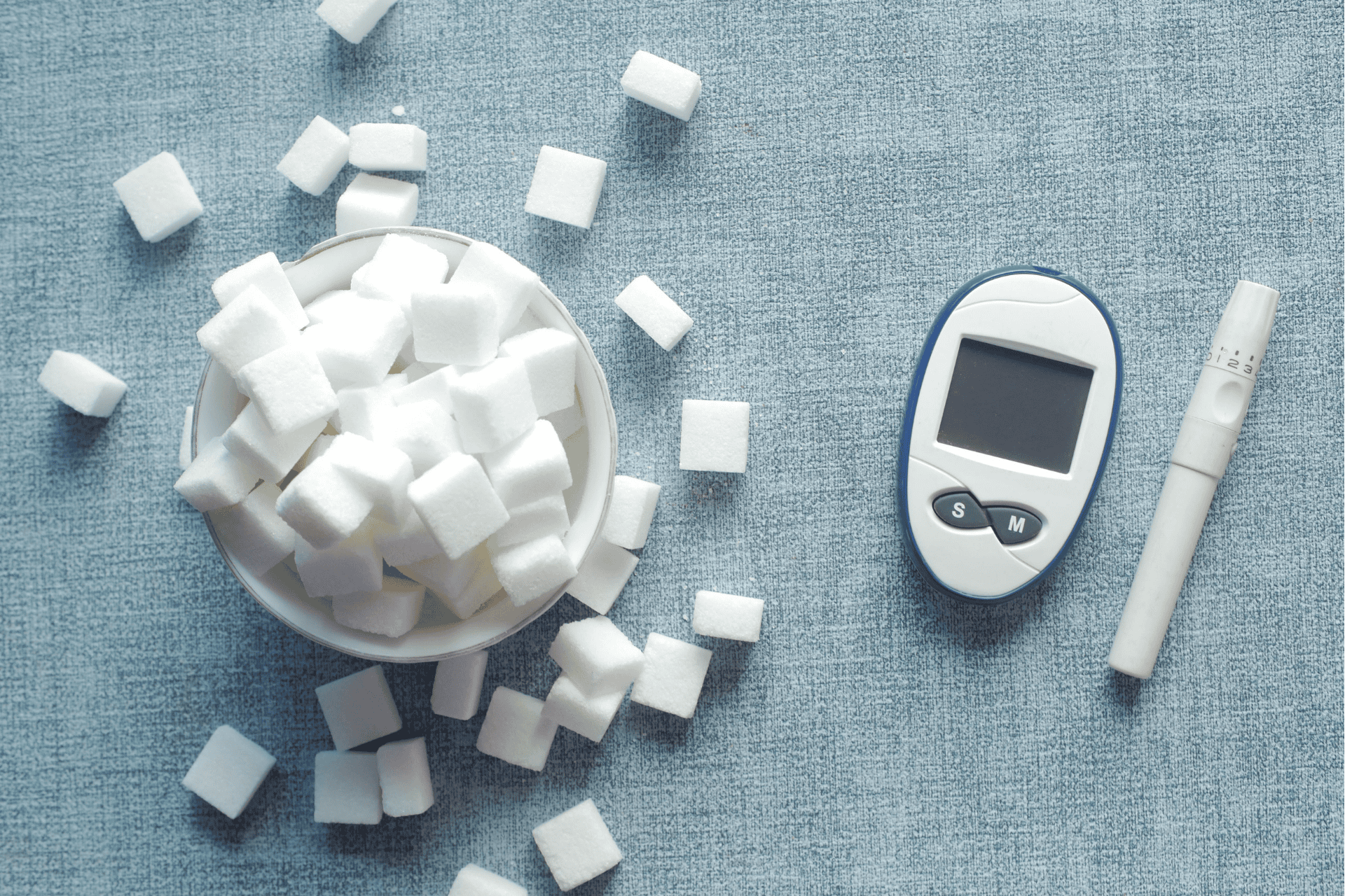 Bowl of sugar cubes next to a glucose meter and lancet on a blue surface.