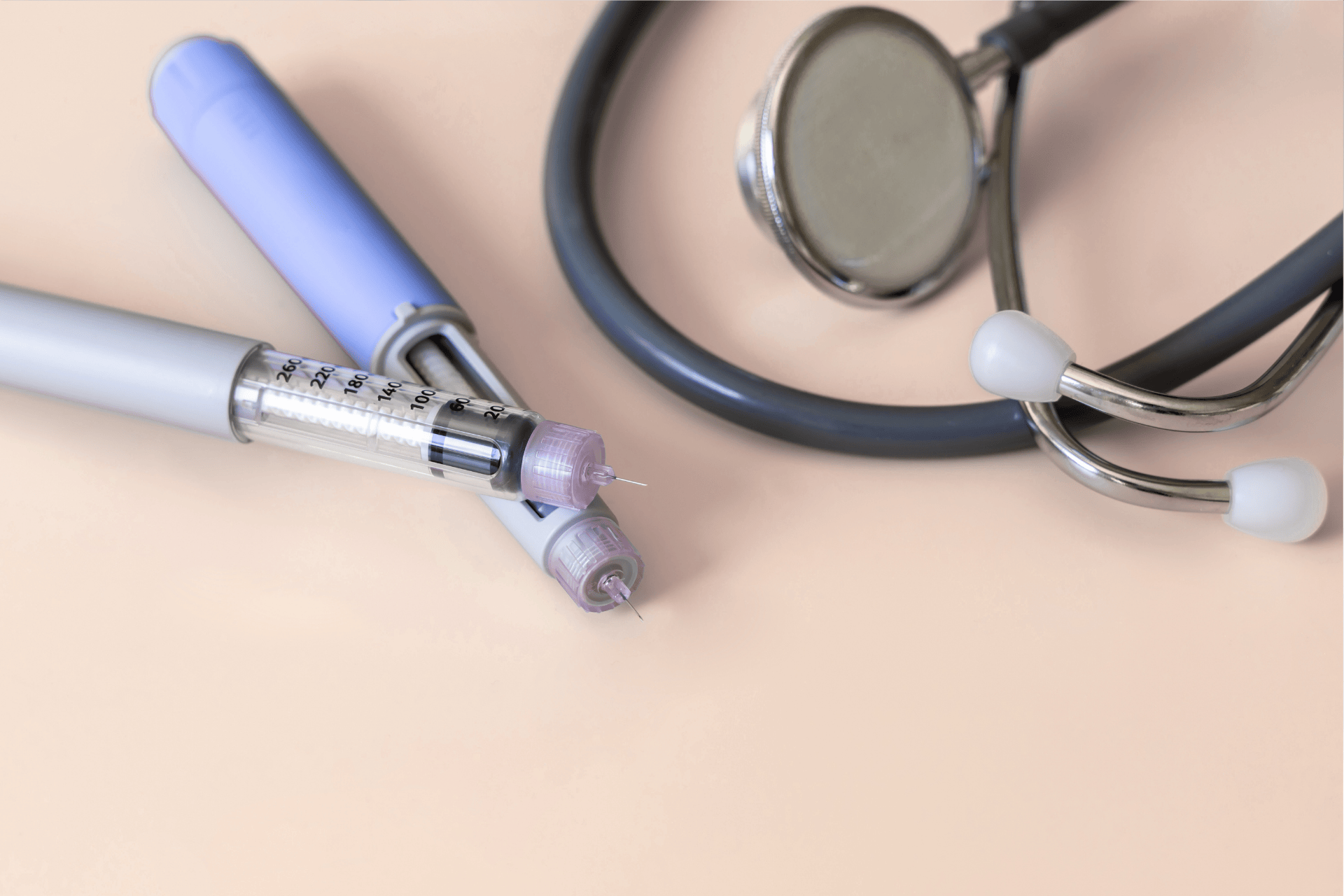 Two insulin pens and a stethoscope placed on a light pink surface