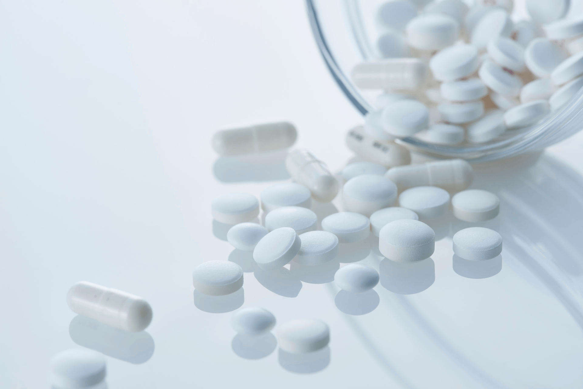 White pills and capsules scattered near a glass container on a reflective surface
