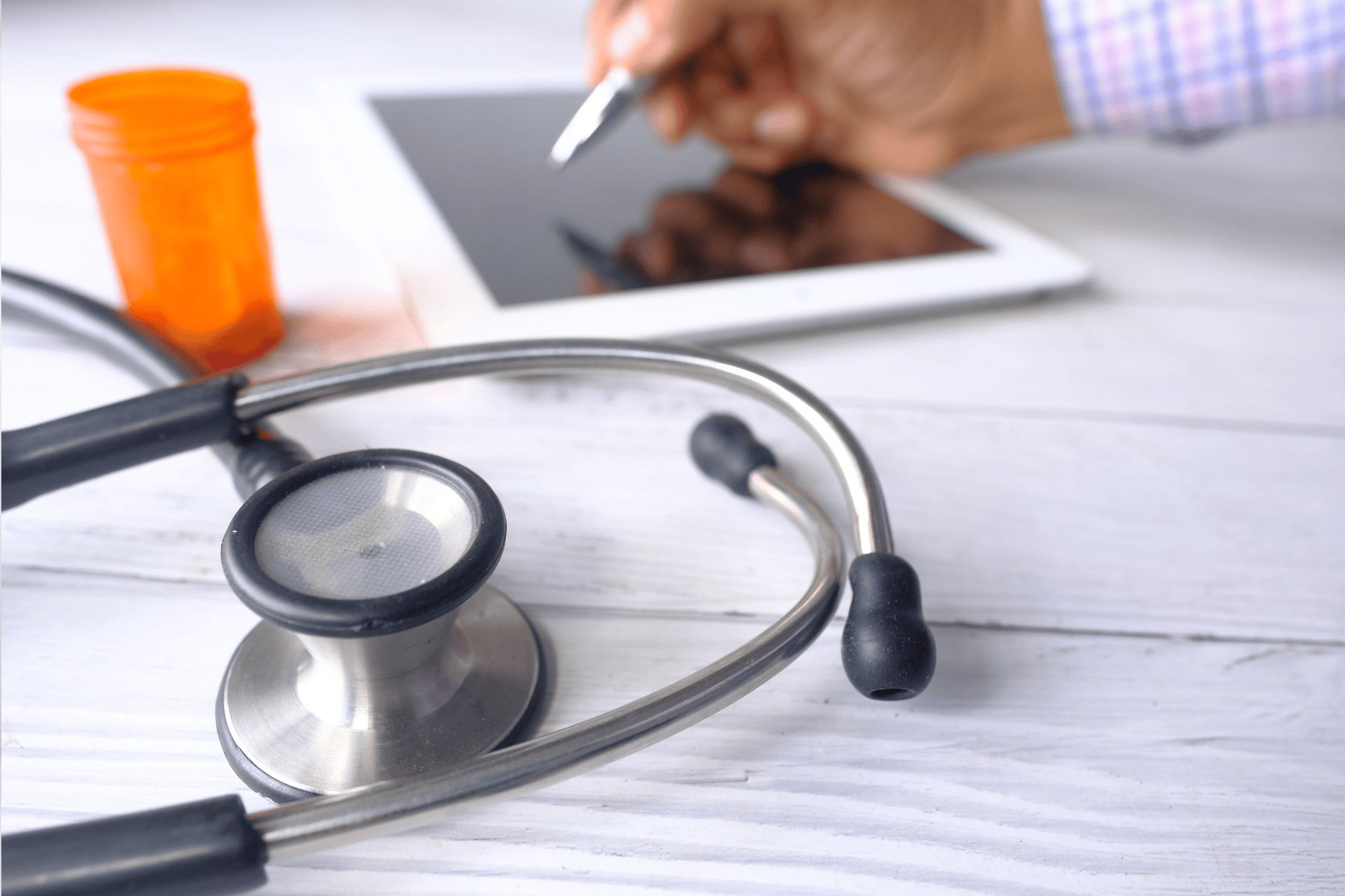 Stethoscope and tablet on a desk with a person using a stylus in the background.