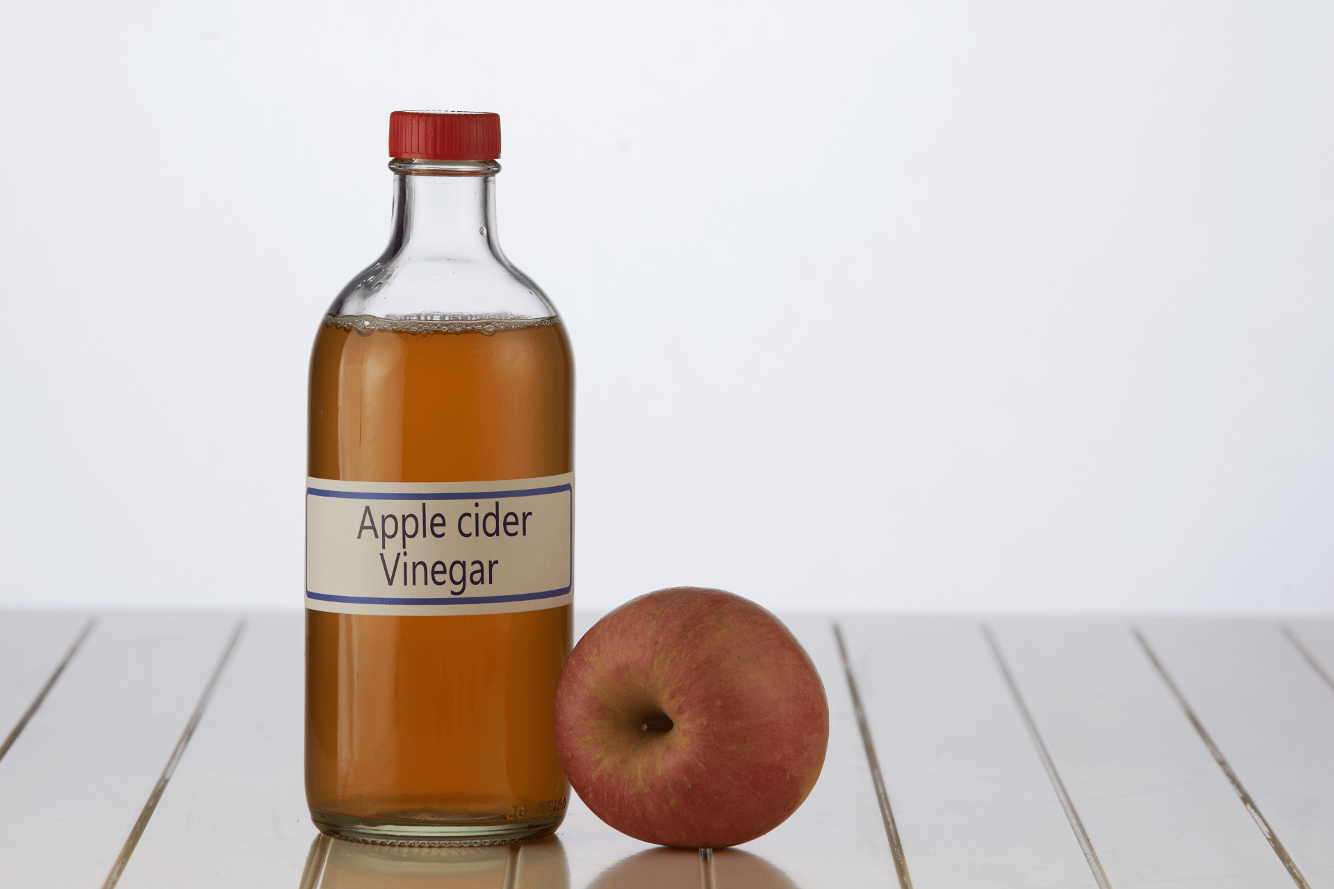 Bottle of apple cider vinegar beside a red apple on a white surface.
