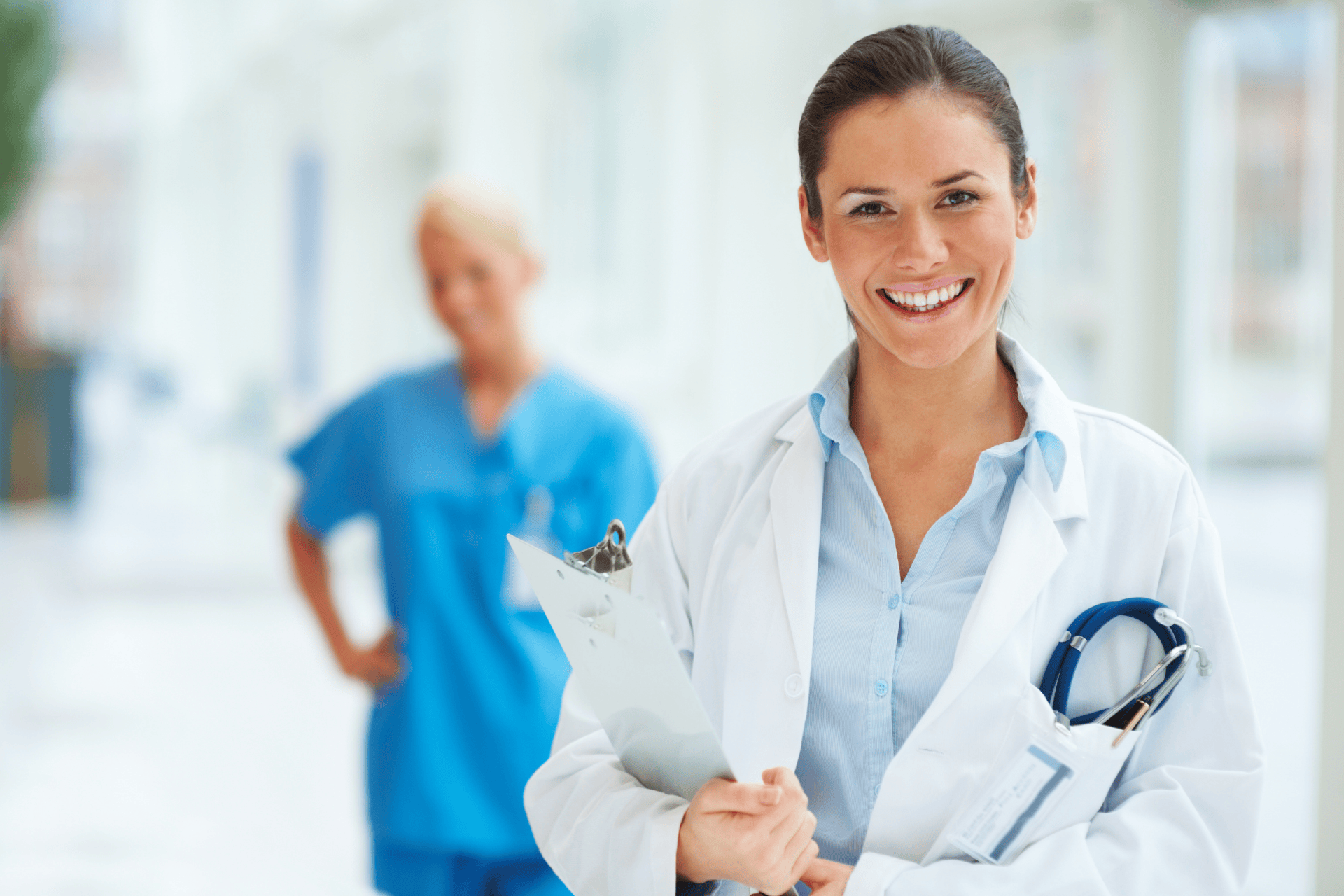 Smiling female doctor holding a clipboard with another healthcare worker in the background.