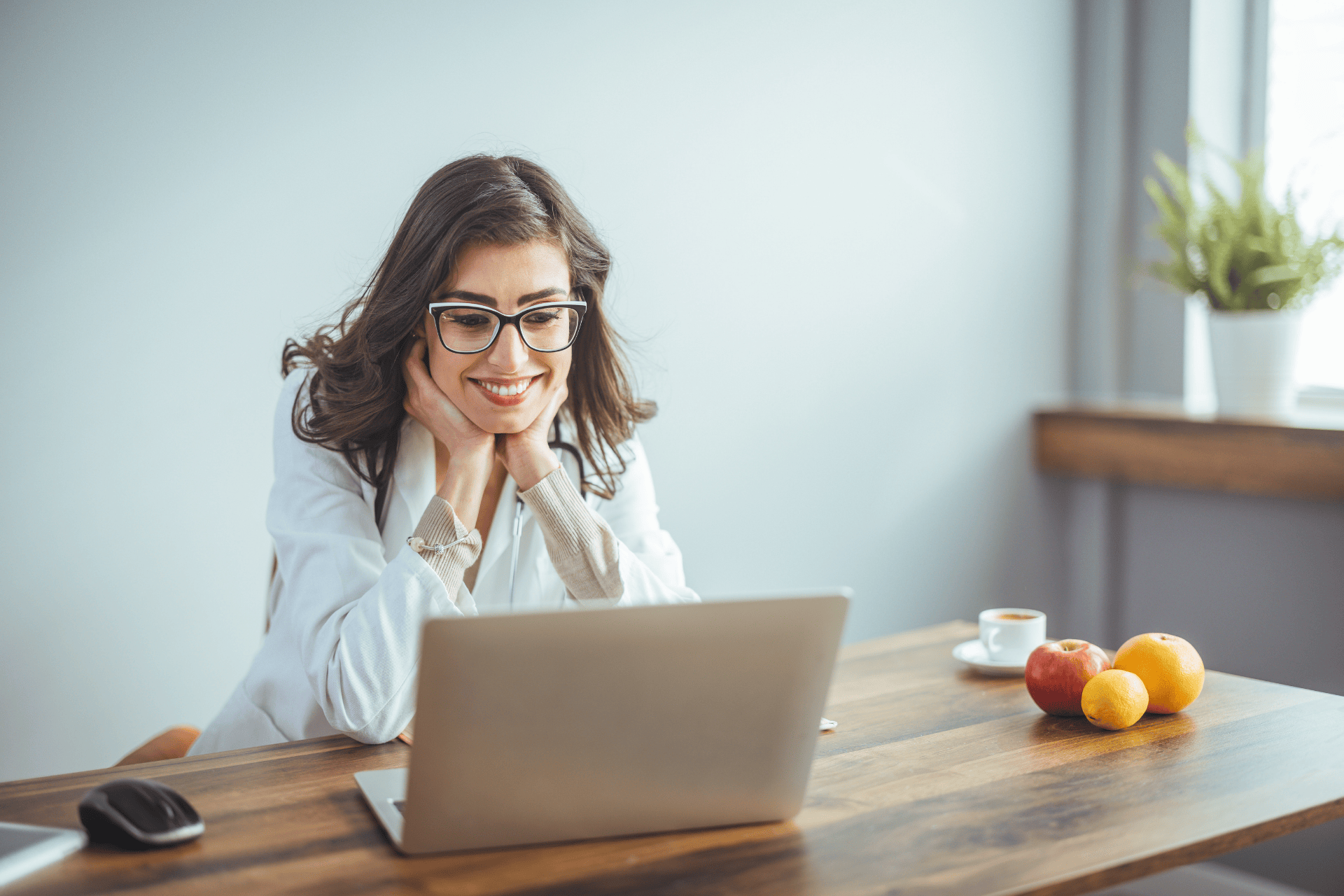 Smiling woman in glasses sitting at a desk using a laptop with fruit and coffee nearby.