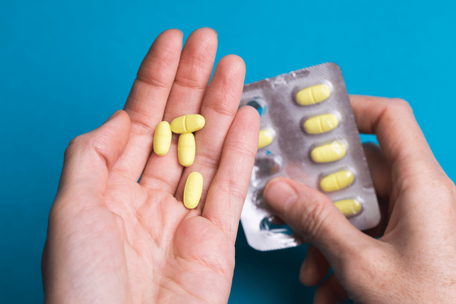 Close-up of hands holding yellow pills and a blister pack against a blue background.