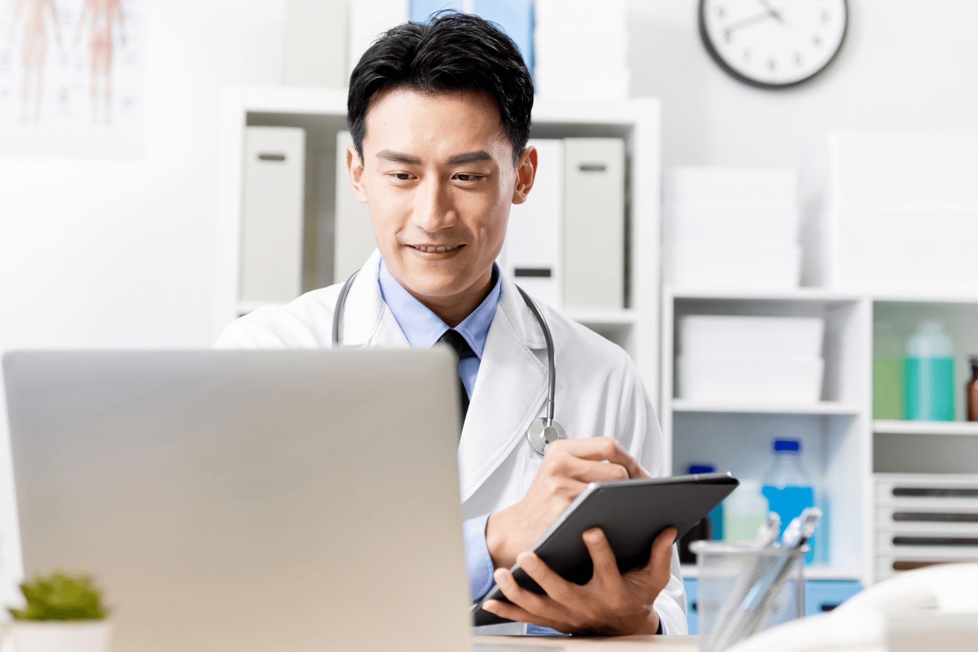 Smiling male doctor with a stethoscope using a tablet and laptop in an office.