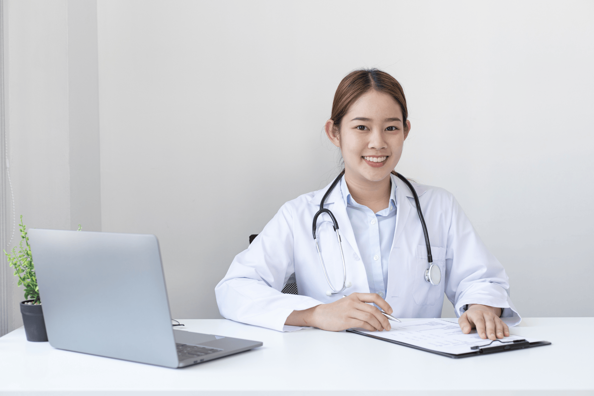 Female doctor sitting at a desk with a laptop, smiling while holding a clipboard.