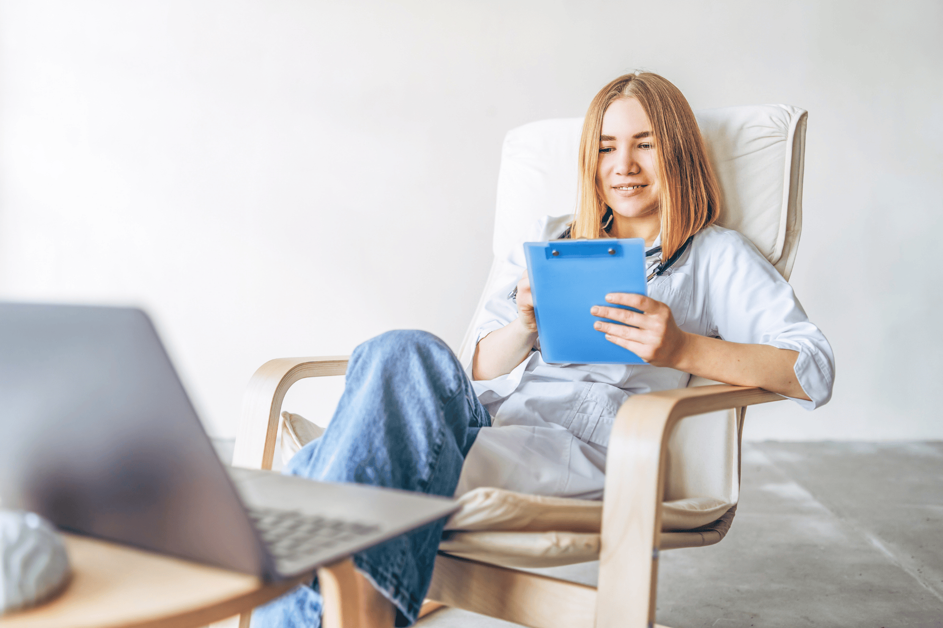Female doctor sitting in a chair, holding a blue clipboard while looking at a laptop.