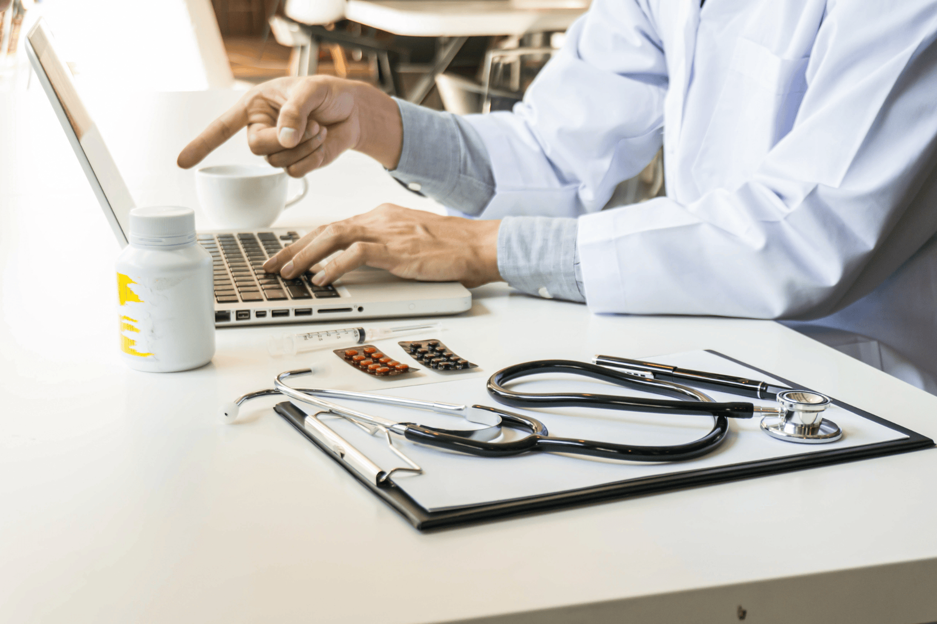 Close-up of a doctor using a laptop with medical tools and medication on the desk.