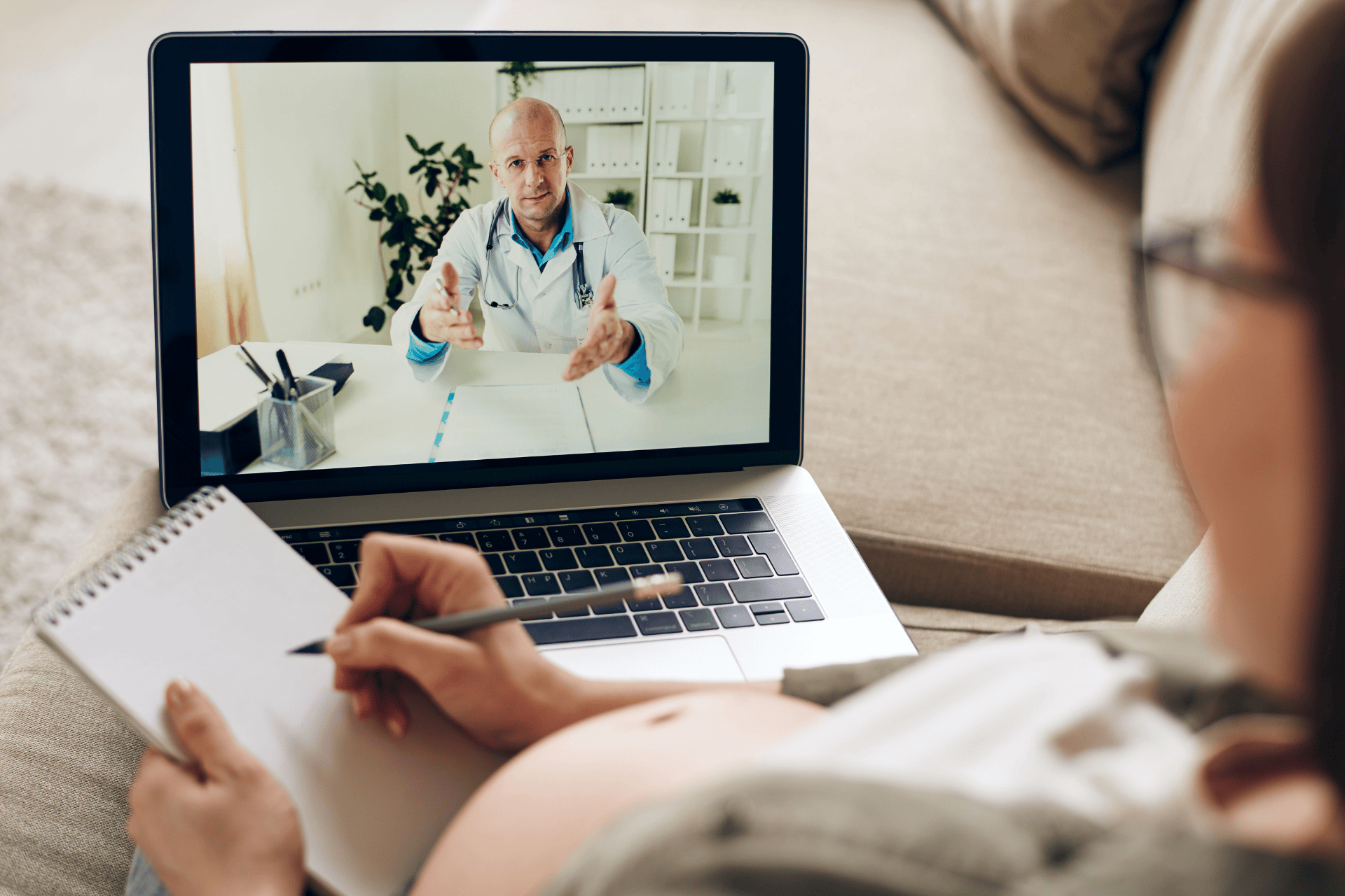 Pregnant woman taking notes during an online video call with a doctor.