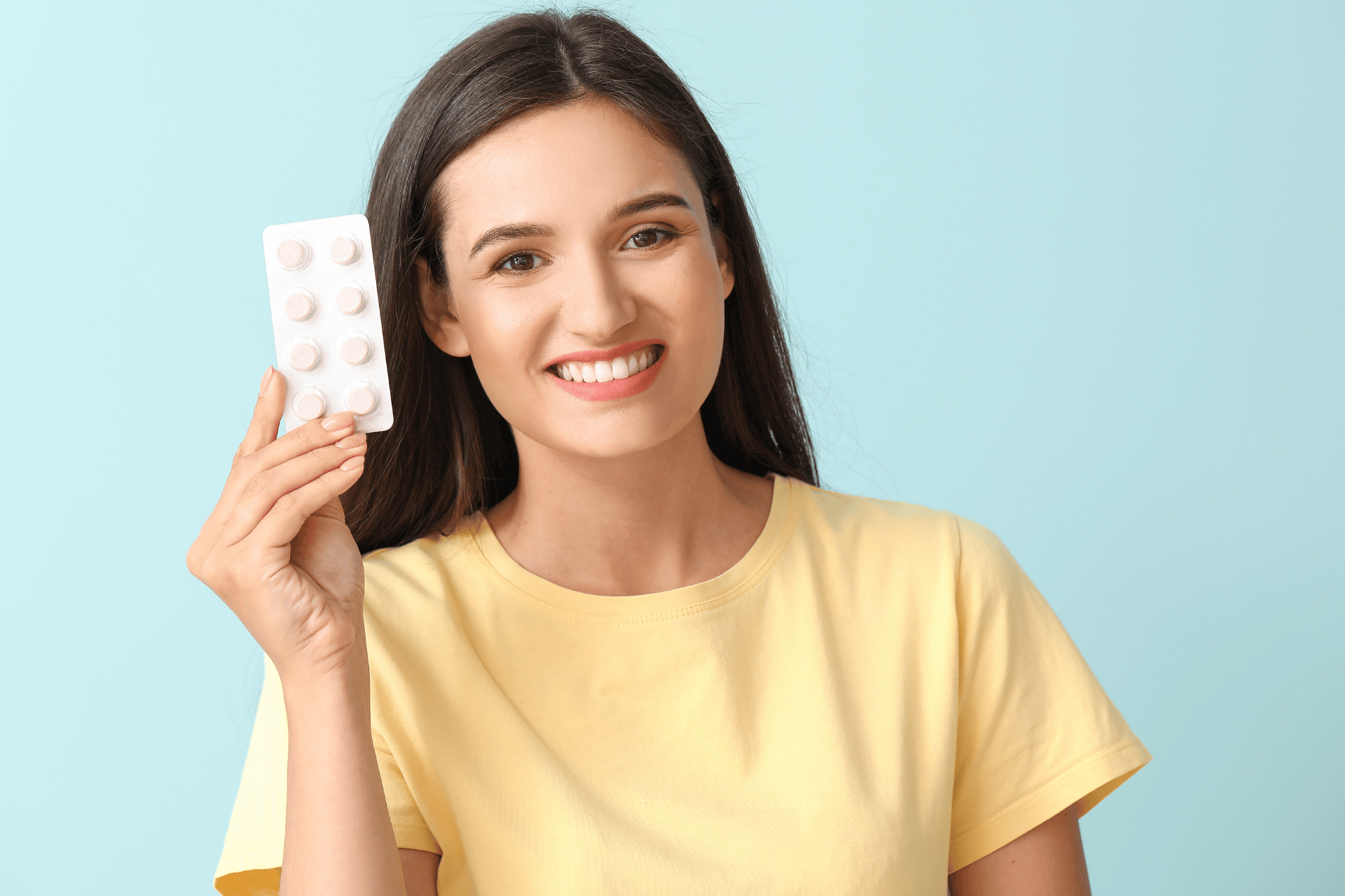 A smiling woman in a yellow shirt holding a blister pack of pills against a light blue background