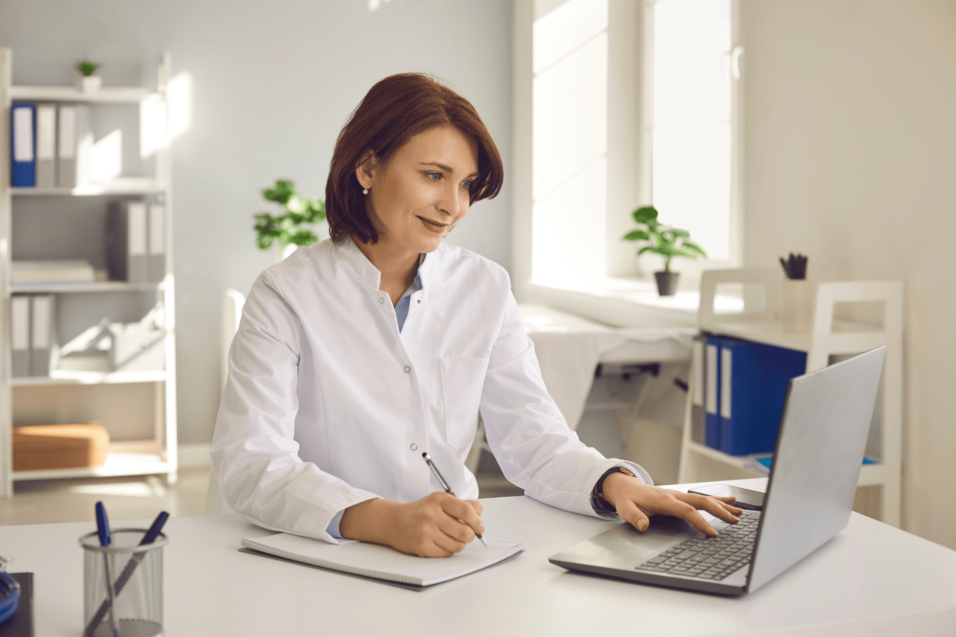 Female doctor writing notes while working on a laptop in a bright medical office.