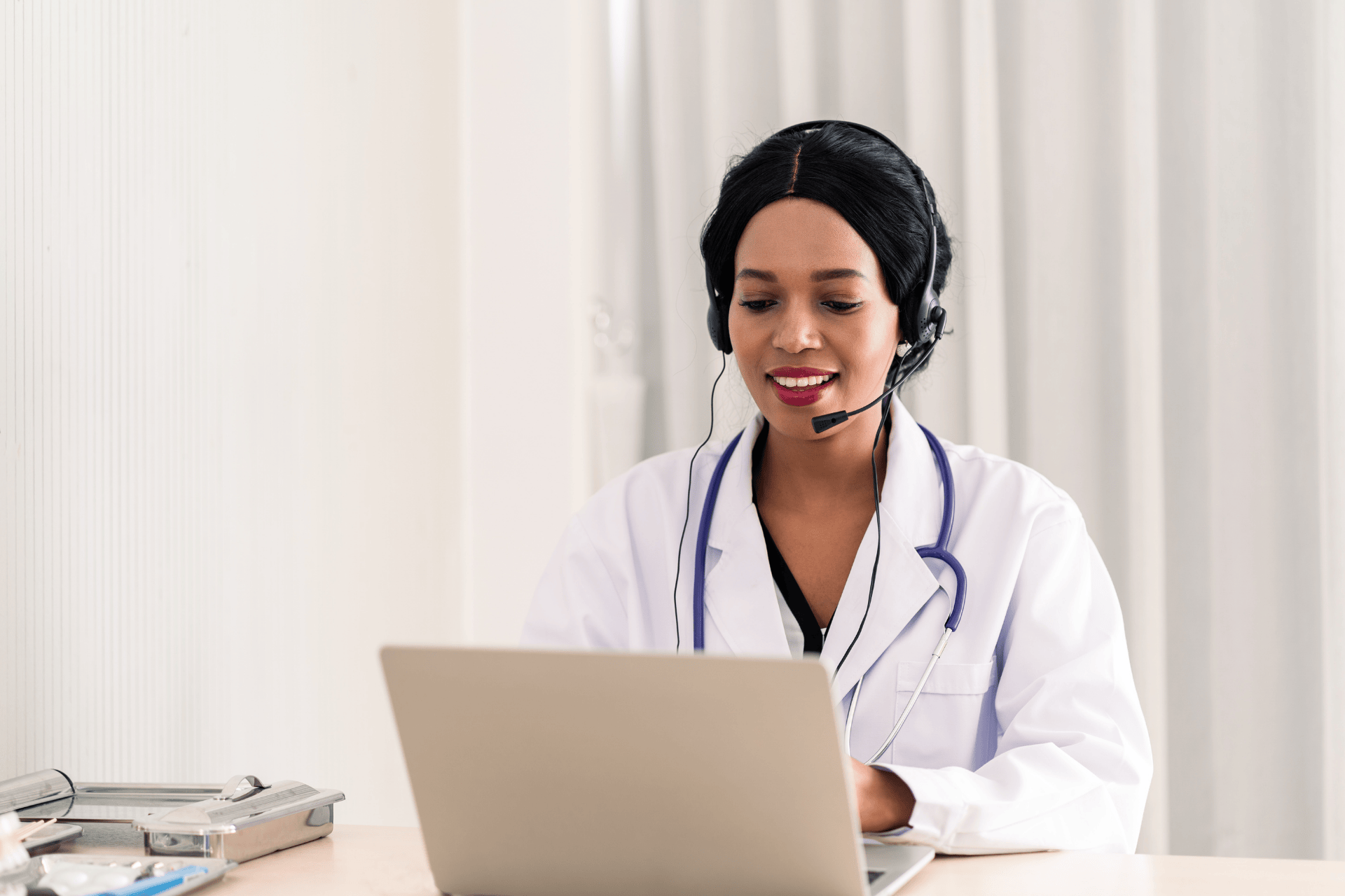 A doctor wearing a headset and white coat speaks while looking at a laptop during a telehealth session.