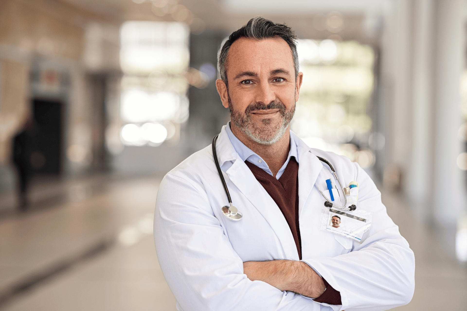 Male doctor in a white coat standing with arms crossed in a bright hospital hallway.