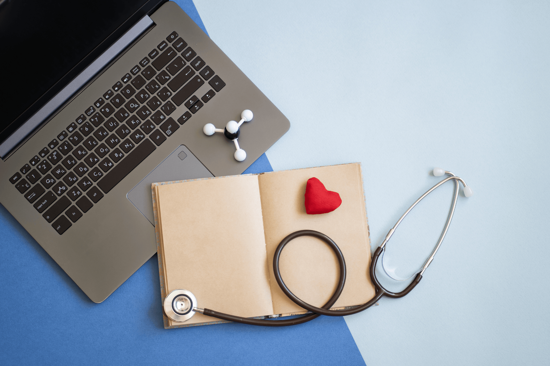 Laptop, open notebook with a small heart, and a stethoscope arranged on a blue background.