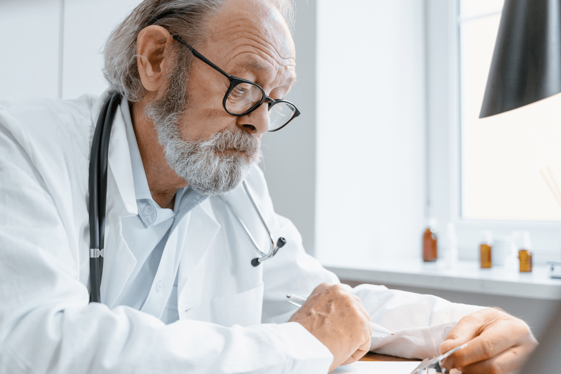 Older male doctor with glasses and a stethoscope writing on a clipboard.