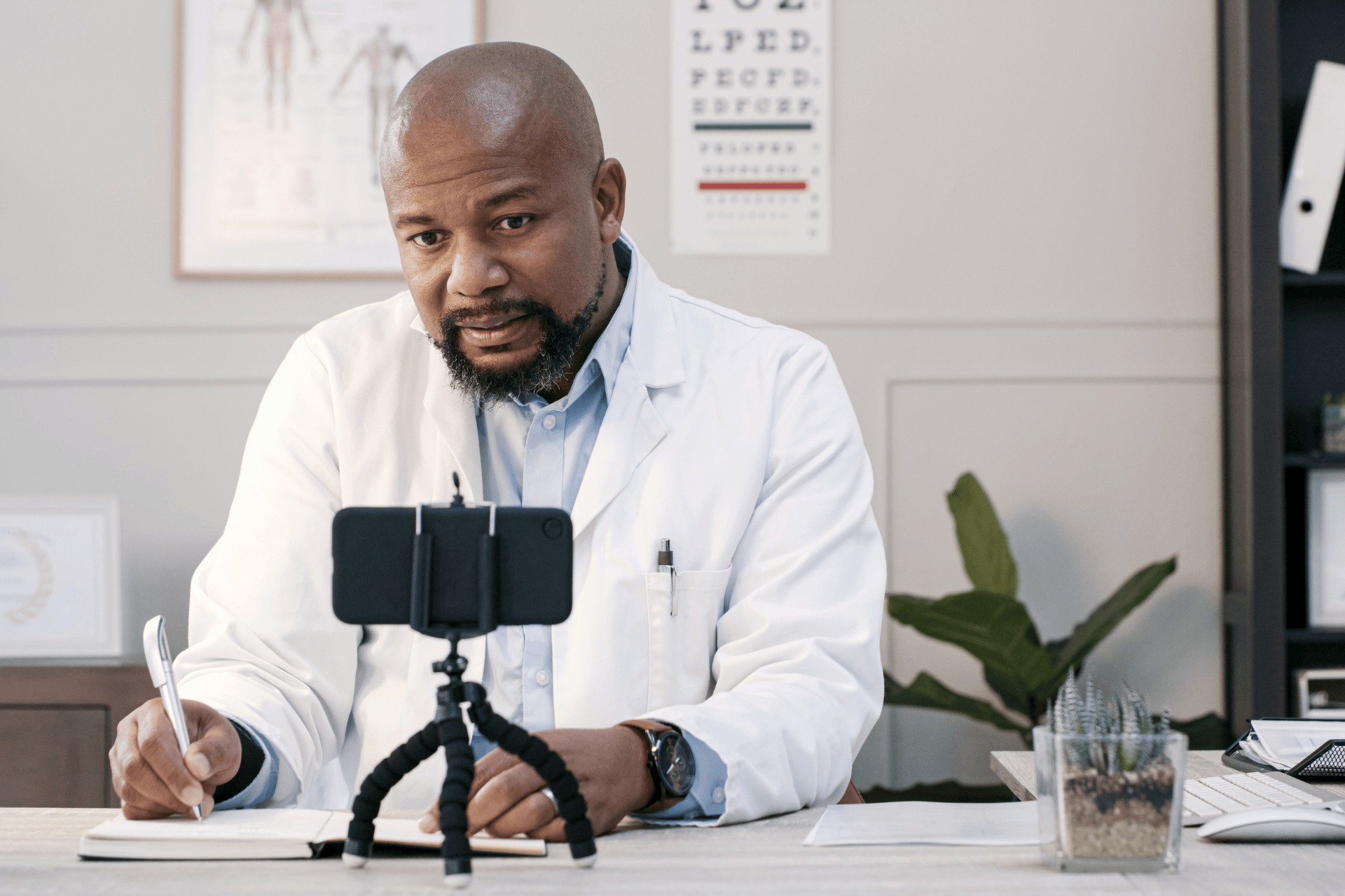 Doctor writing notes while speaking to a smartphone on a tripod during a virtual visit.