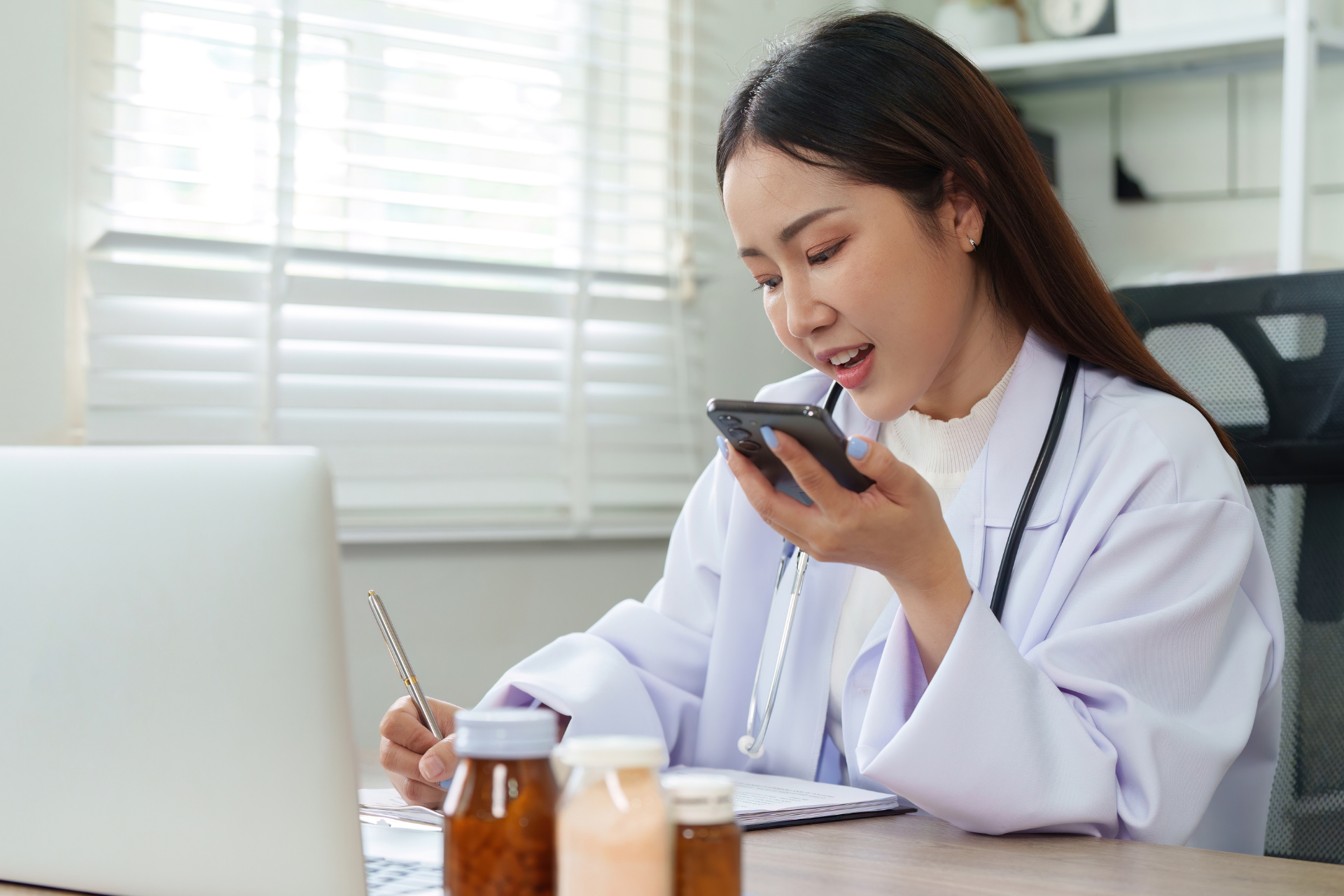 Doctor speaking into a smartphone while writing notes at her desk.
