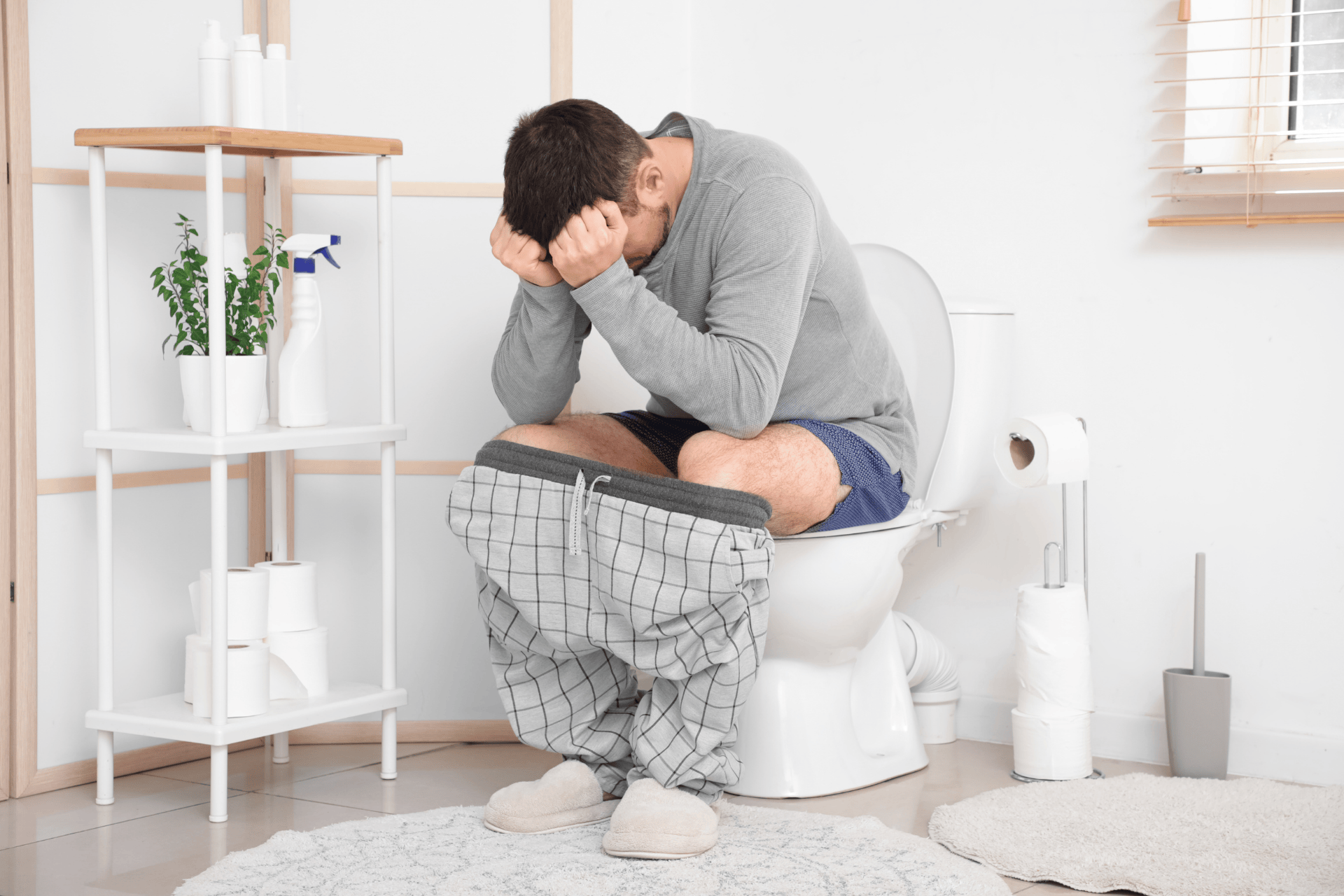 Man sitting on a toilet with his head in his hands, appearing to be in pain or discomfort.