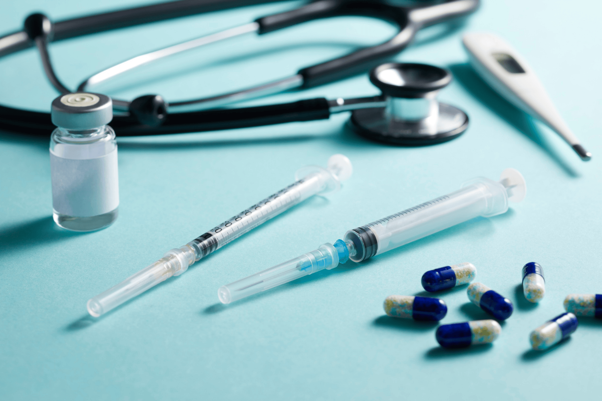Close-up of syringes, capsules, a vial, a stethoscope, and a thermometer on a blue surface.