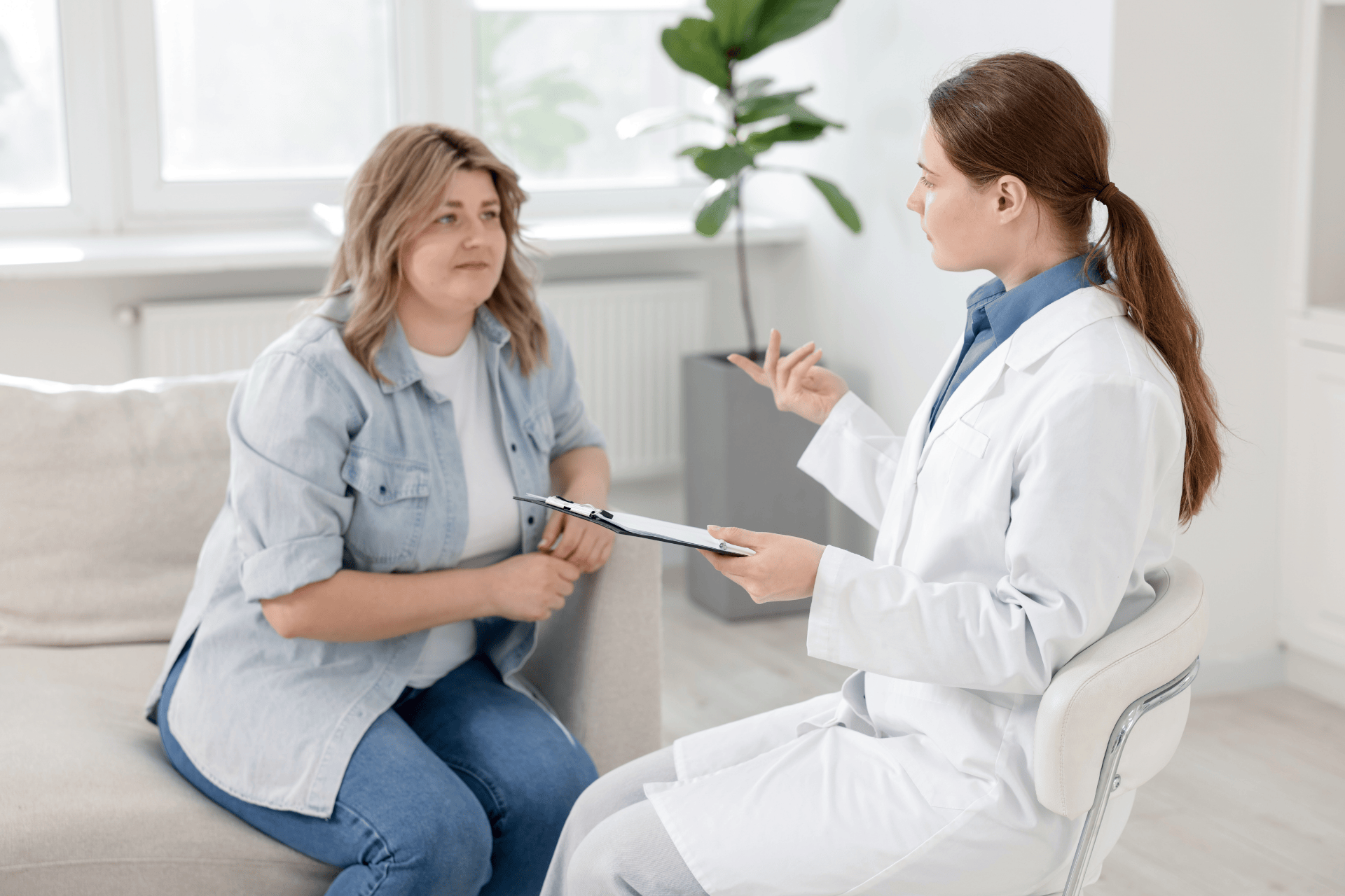 Doctor speaking with a patient during an in-person consultation, holding a clipboard while the patient listens.