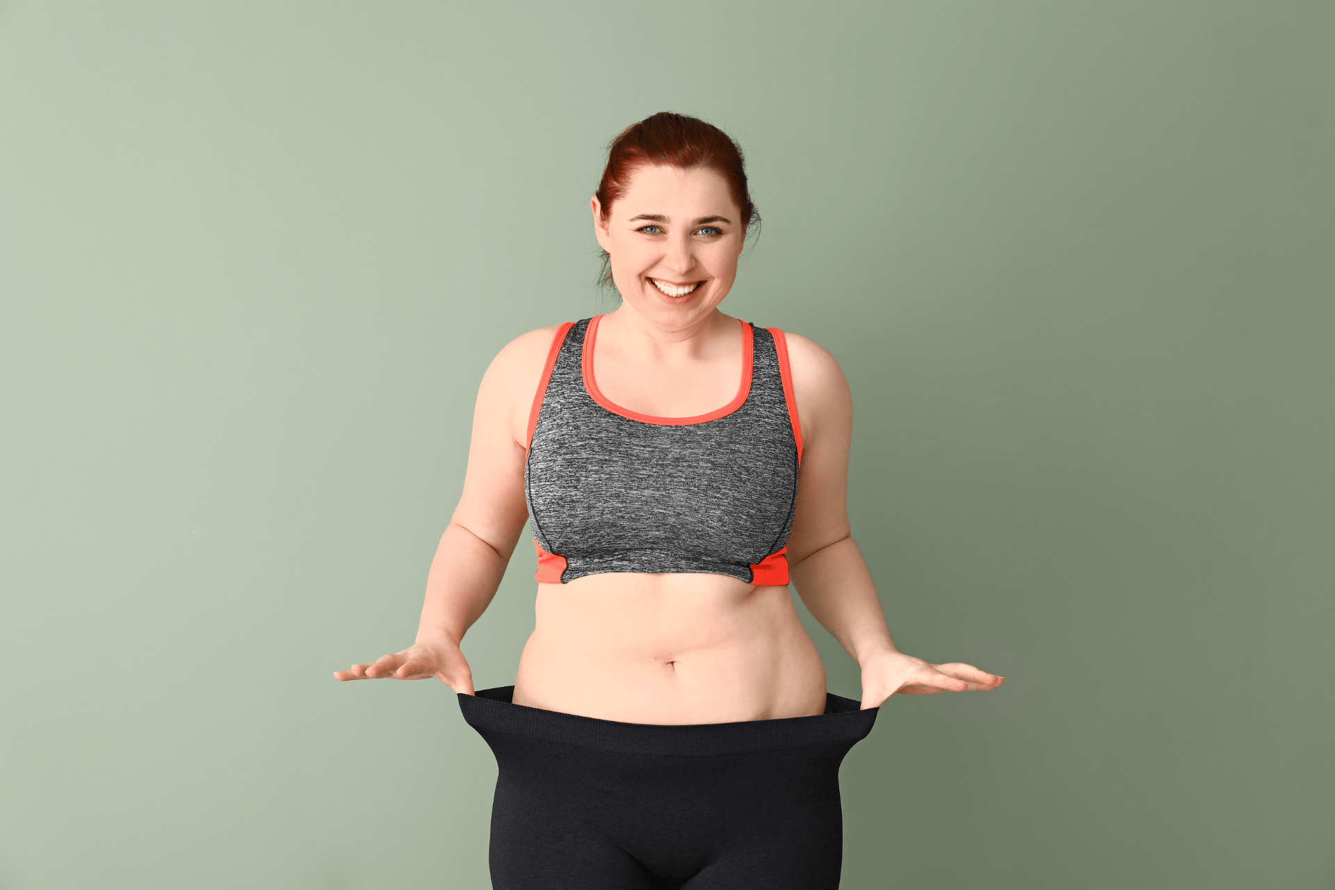 Smiling woman in workout clothes showing loose pants after weight loss.