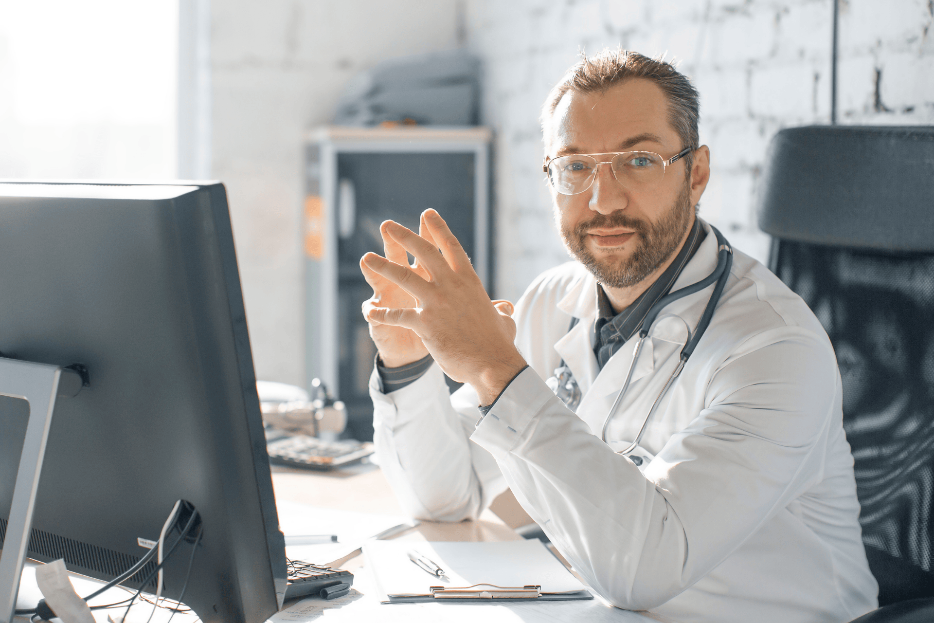Male doctor in a white coat sitting at a desk, looking at the camera with hands clasped.