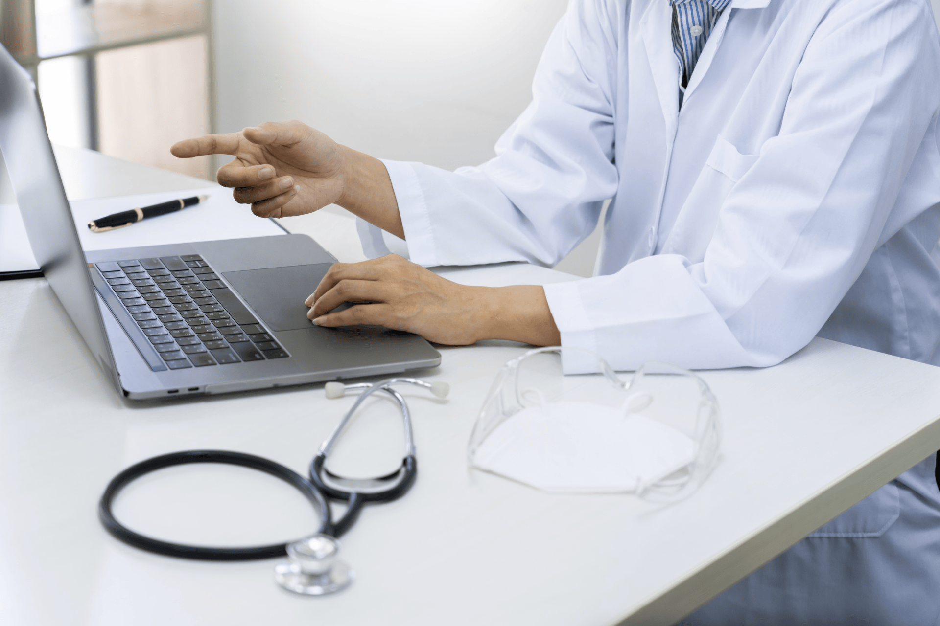 Doctor using a laptop while pointing at the screen, with a stethoscope on the desk.