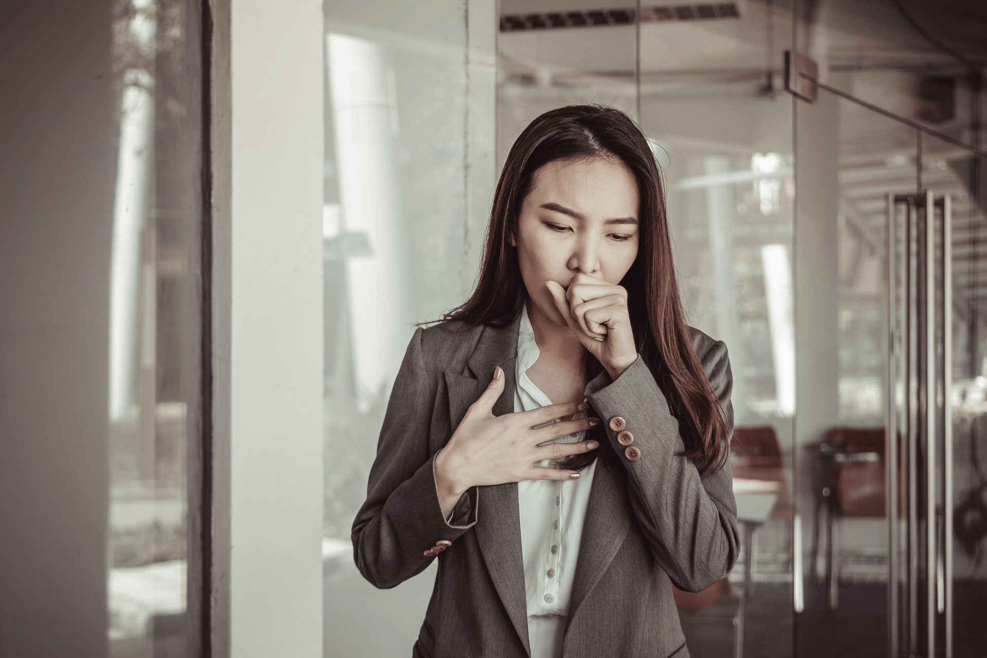 Woman in a business suit coughing and holding her chest in an office setting.