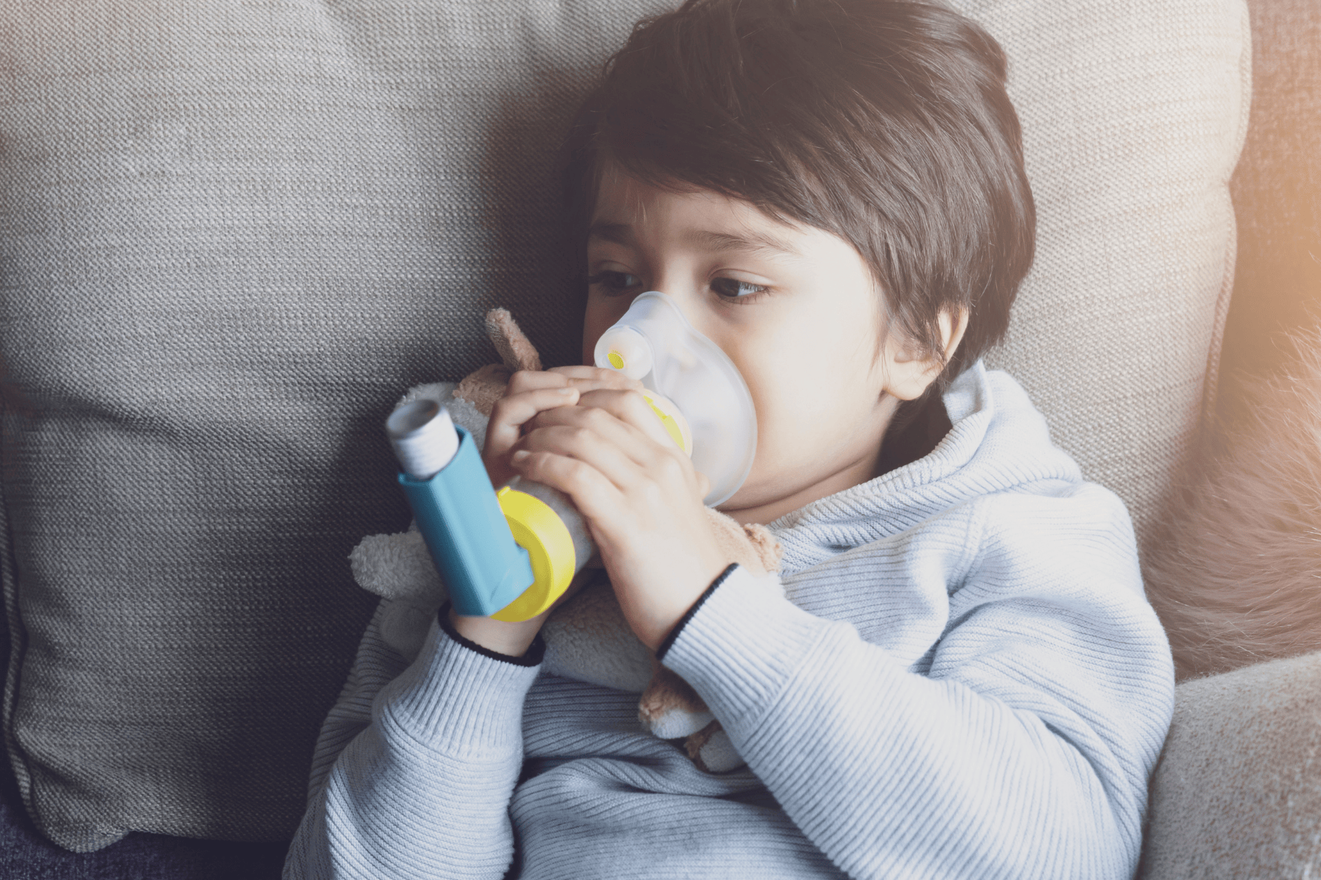 Young child using an inhaler with a spacer mask while lying on a couch.