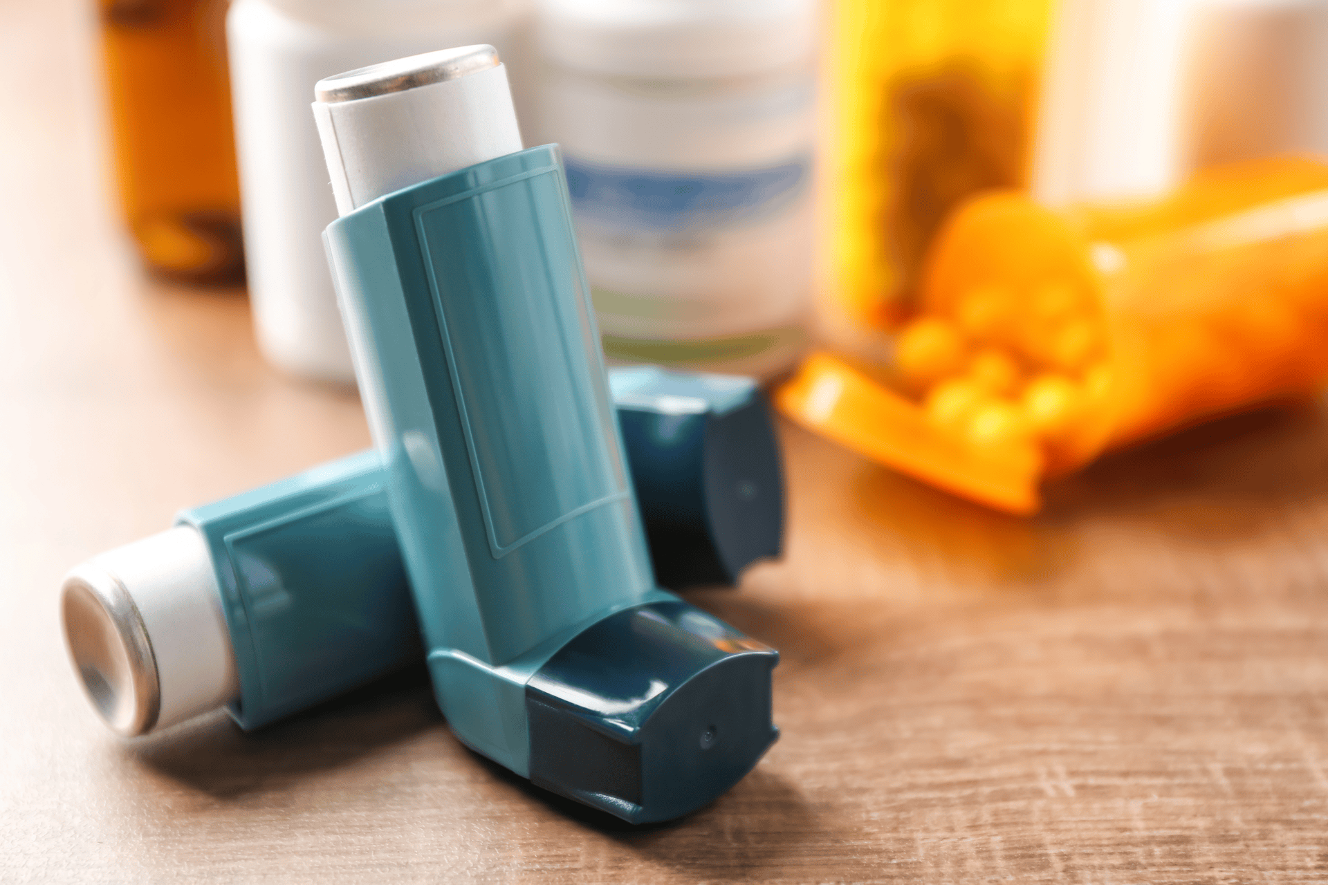 Two asthma inhalers on a table with prescription bottles in the background.
