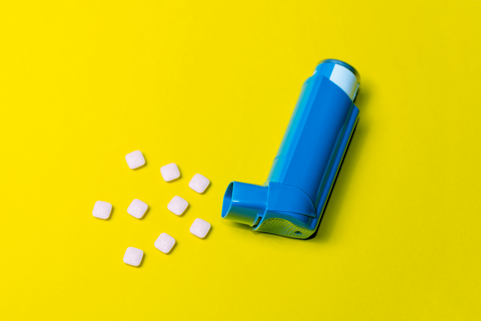 Blue asthma inhaler next to small white pills on a bright yellow background.