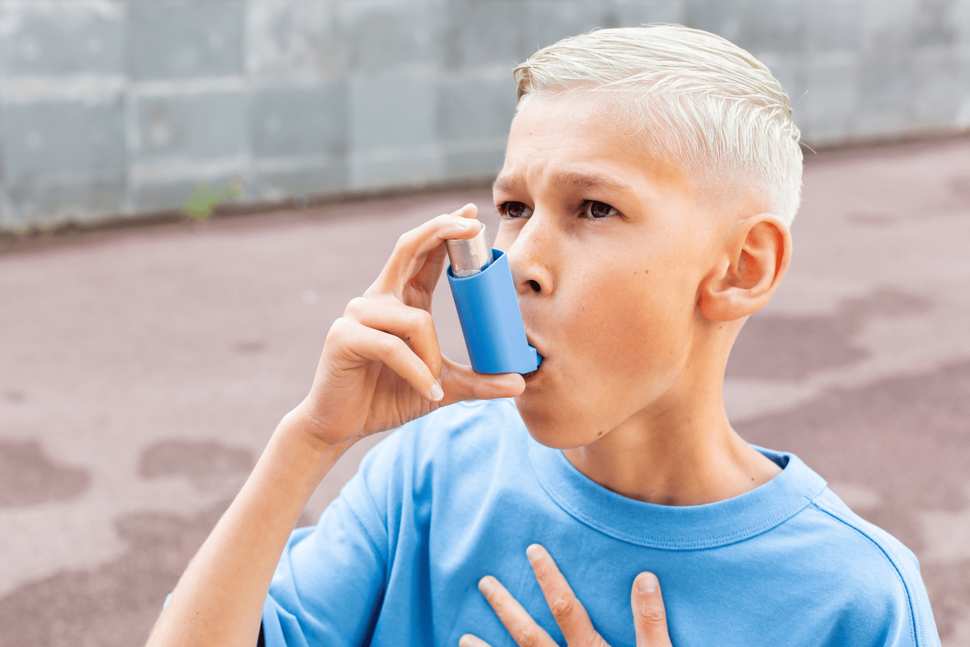 Young boy using a blue asthma inhaler outdoors with his hand on his chest.