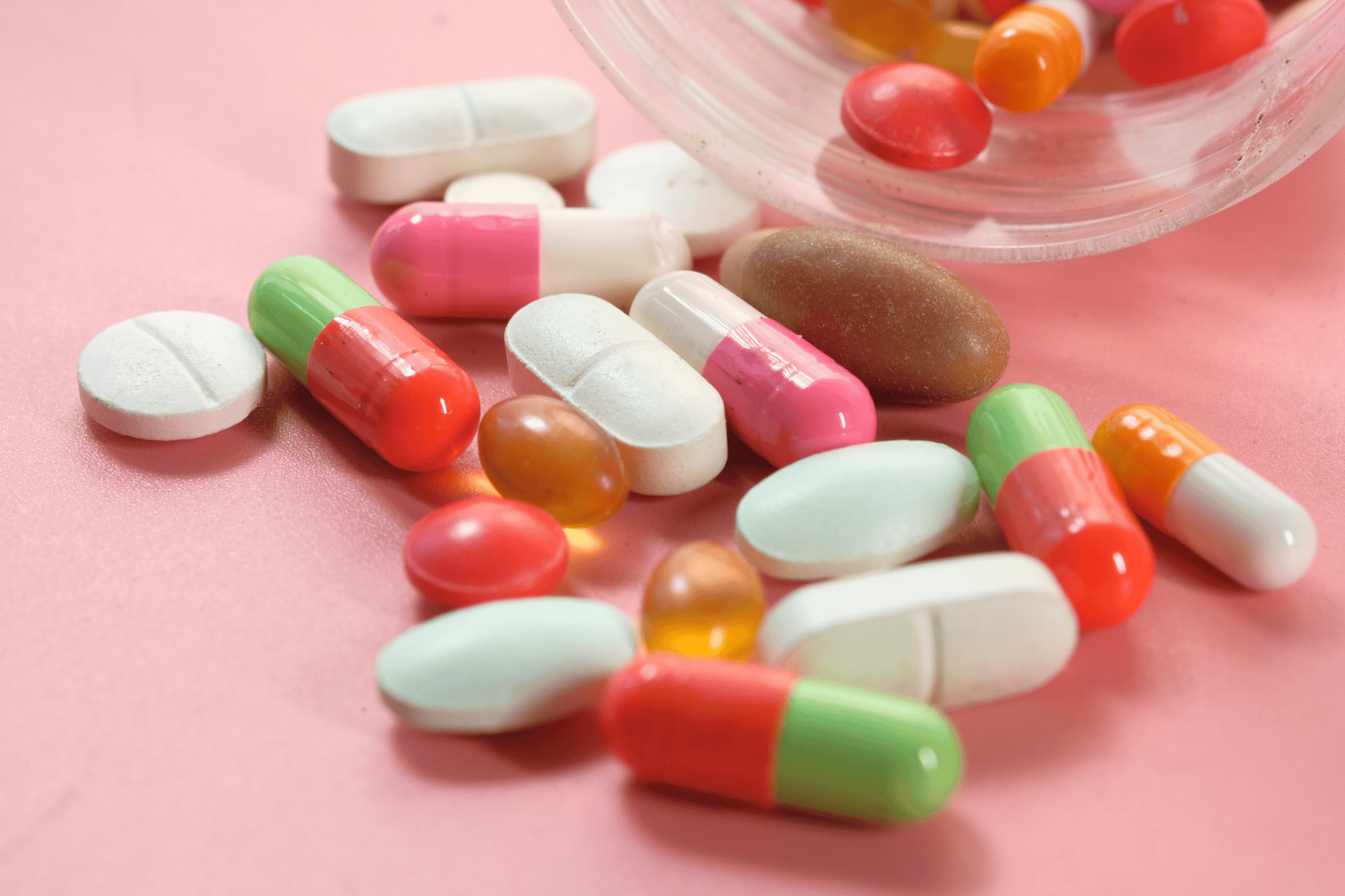 Colorful mix of capsules and tablets spilled from a container onto a pink surface.