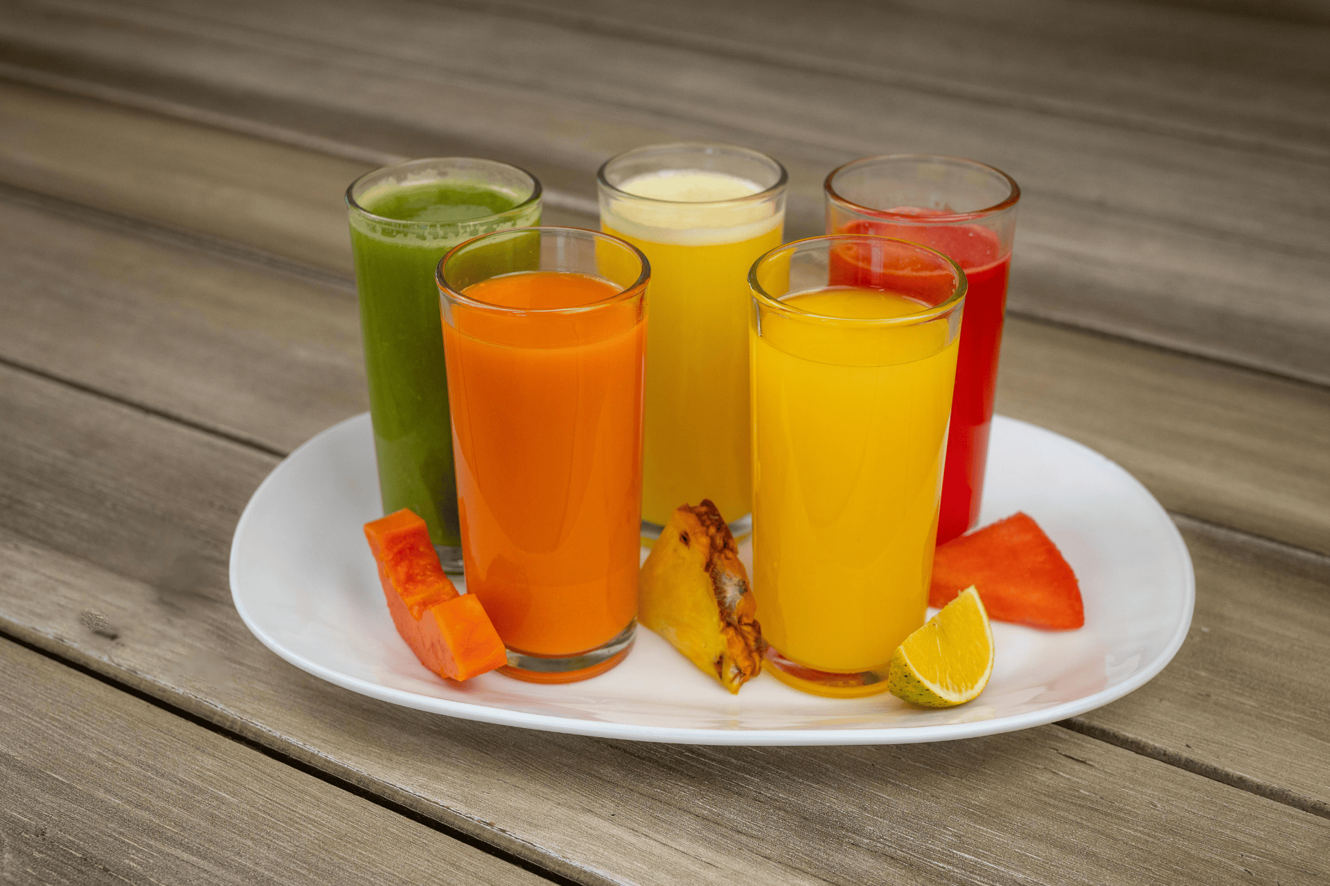 Glasses of colorful fruit and vegetable juices on a plate with fresh fruit pieces.
