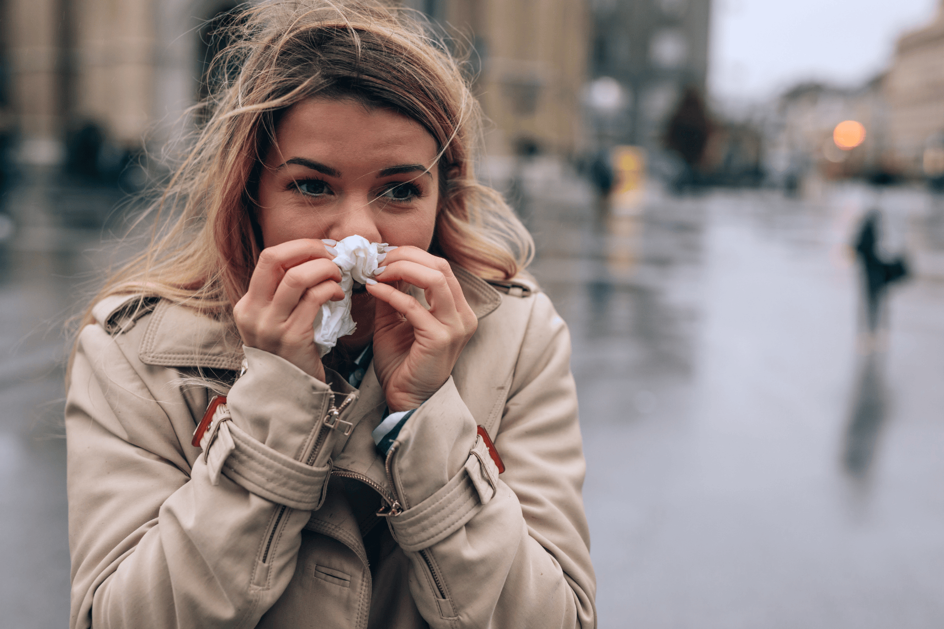 Woman outdoors on a cold, rainy day holding a tissue to her nose as if she has a cold.