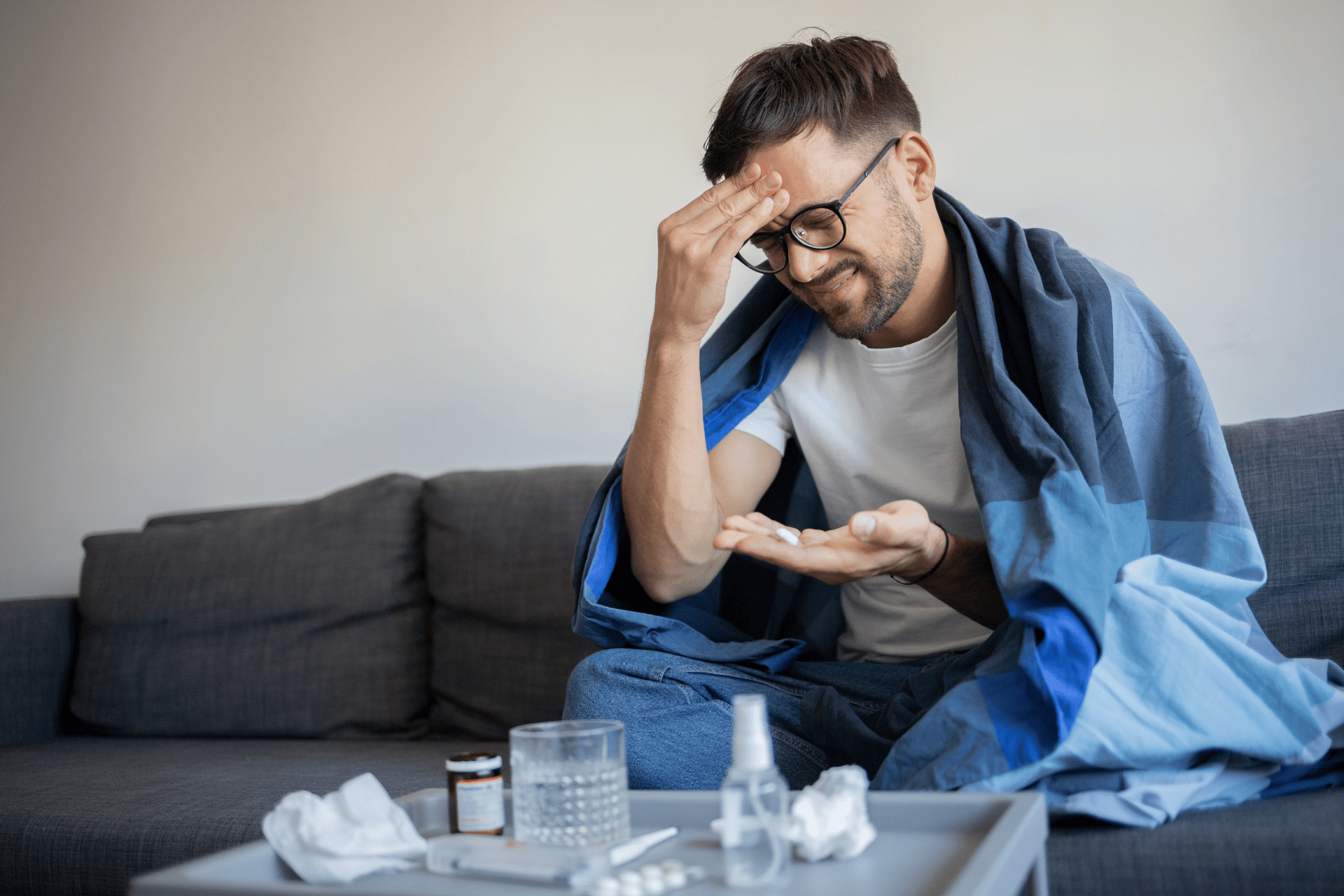 A Man sitting on a couch with a blanket around him, holding his forehead in pain while looking at pills in his hand.