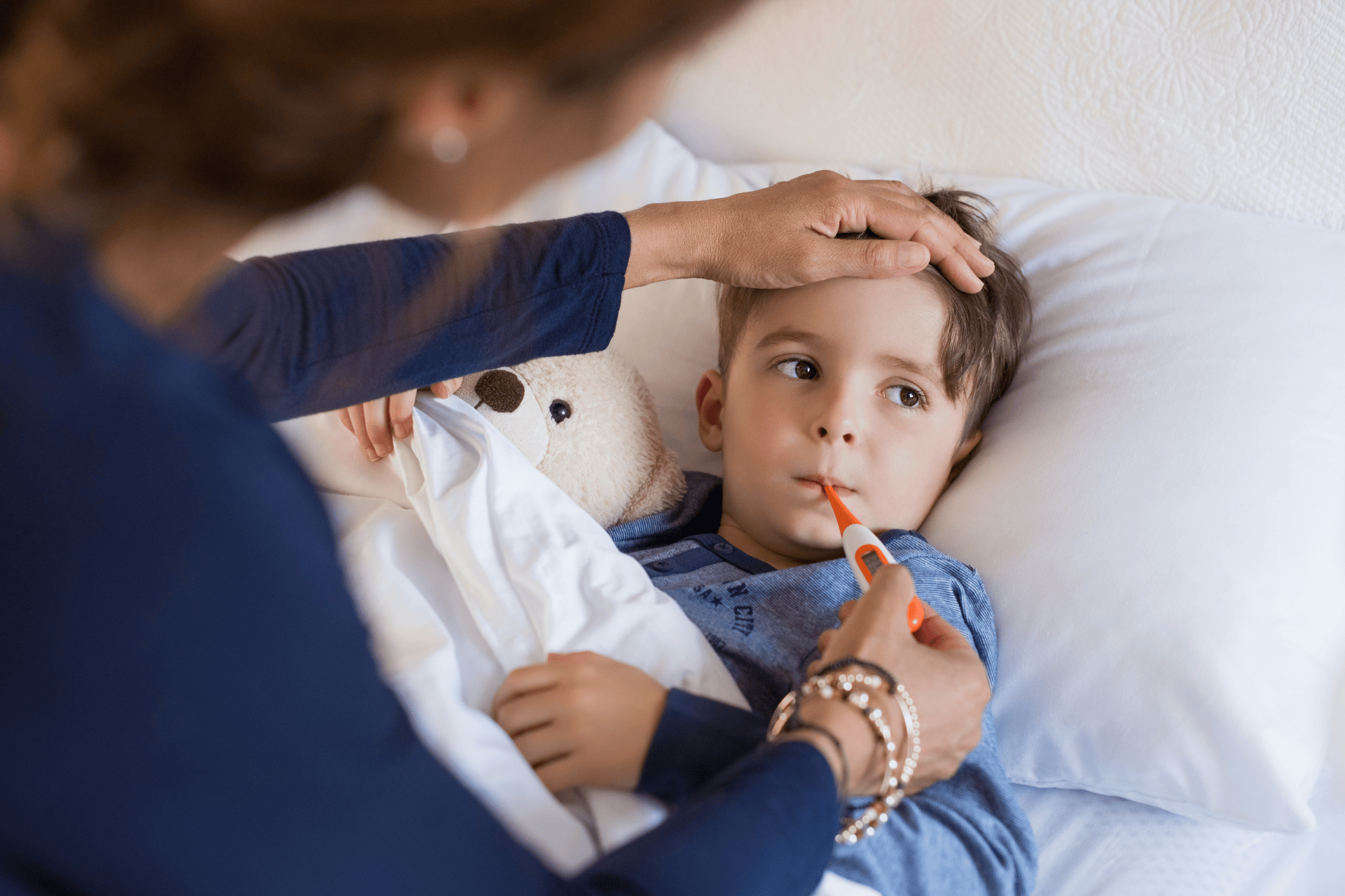 Sick child lying in bed with a thermometer in his mouth while an adult checks his forehead.