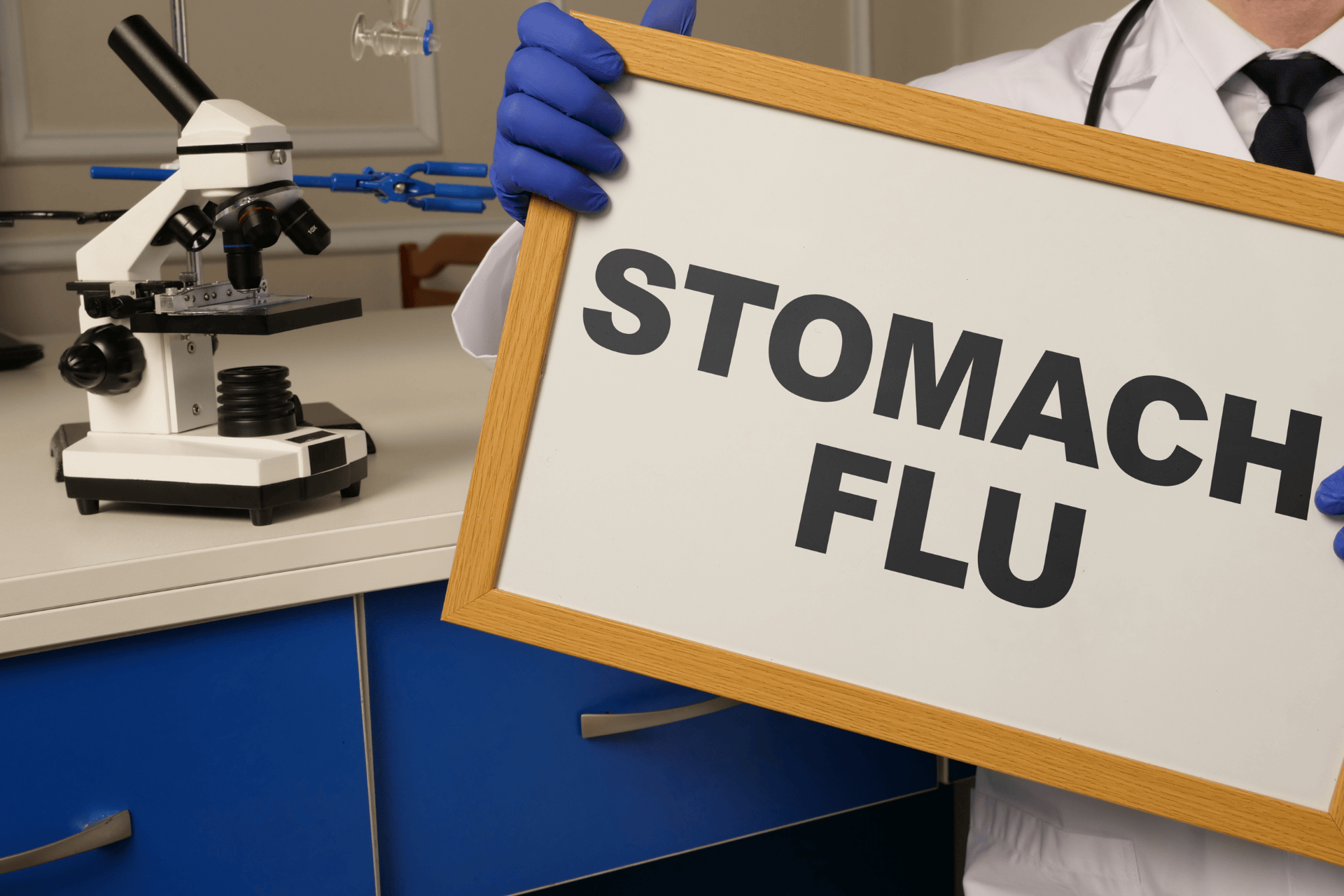 Medical professional holding a sign that reads “STOMACH FLU” next to a microscope in a lab setting.