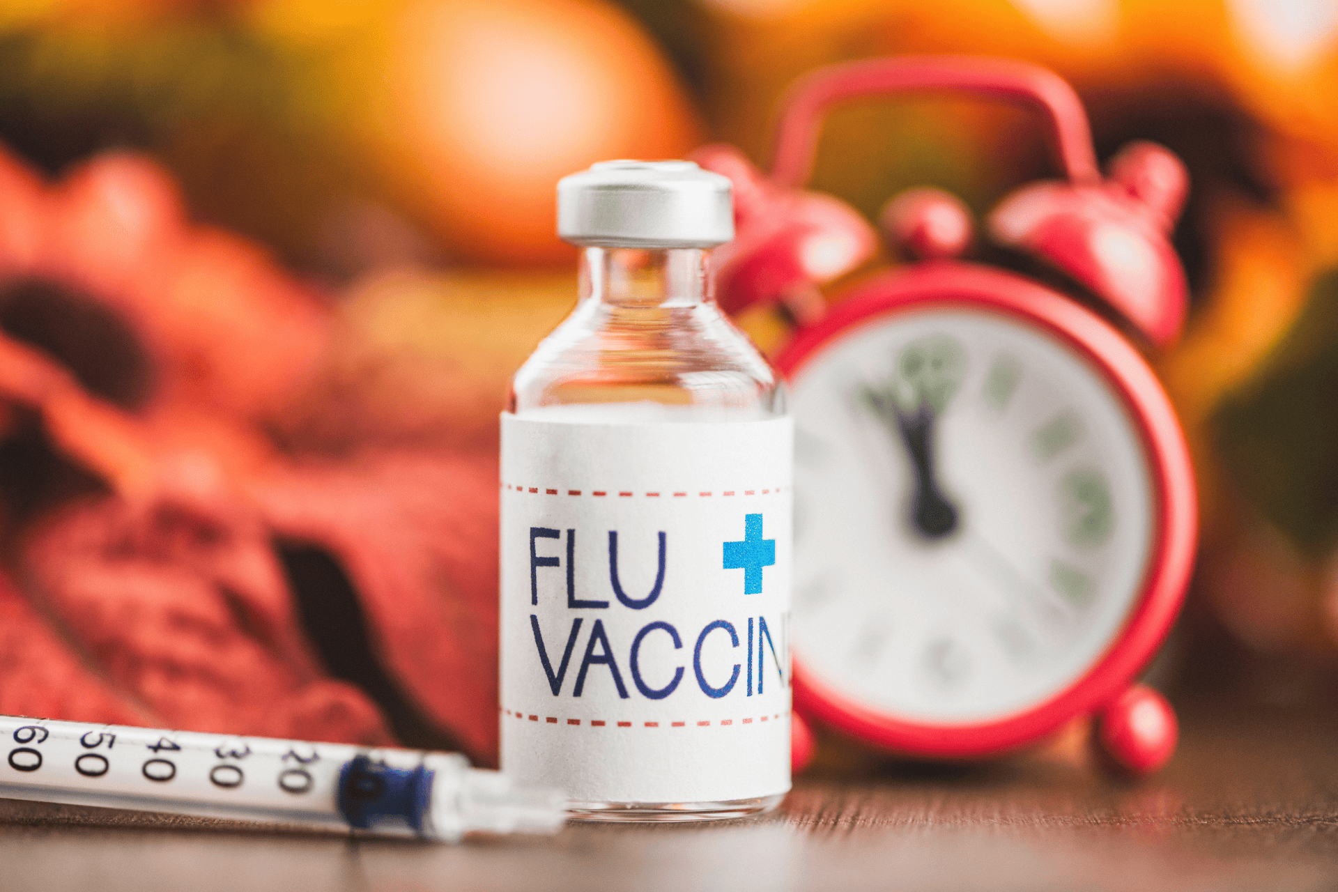 Flu vaccine vial with a syringe beside it and a red alarm clock in the blurred background.