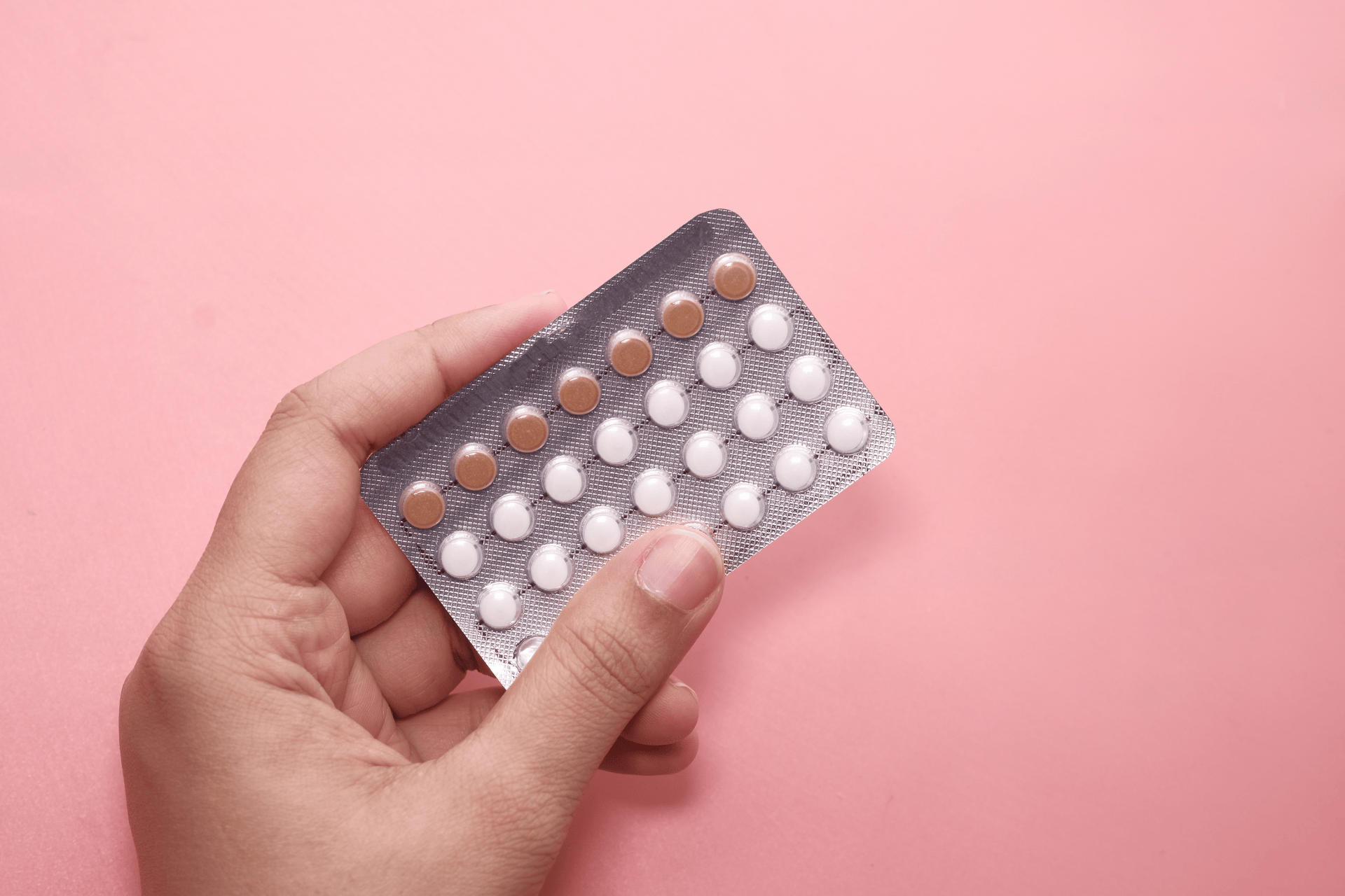 Hand holding a blister pack of birth control pills with white and brown tablets against a pink background.